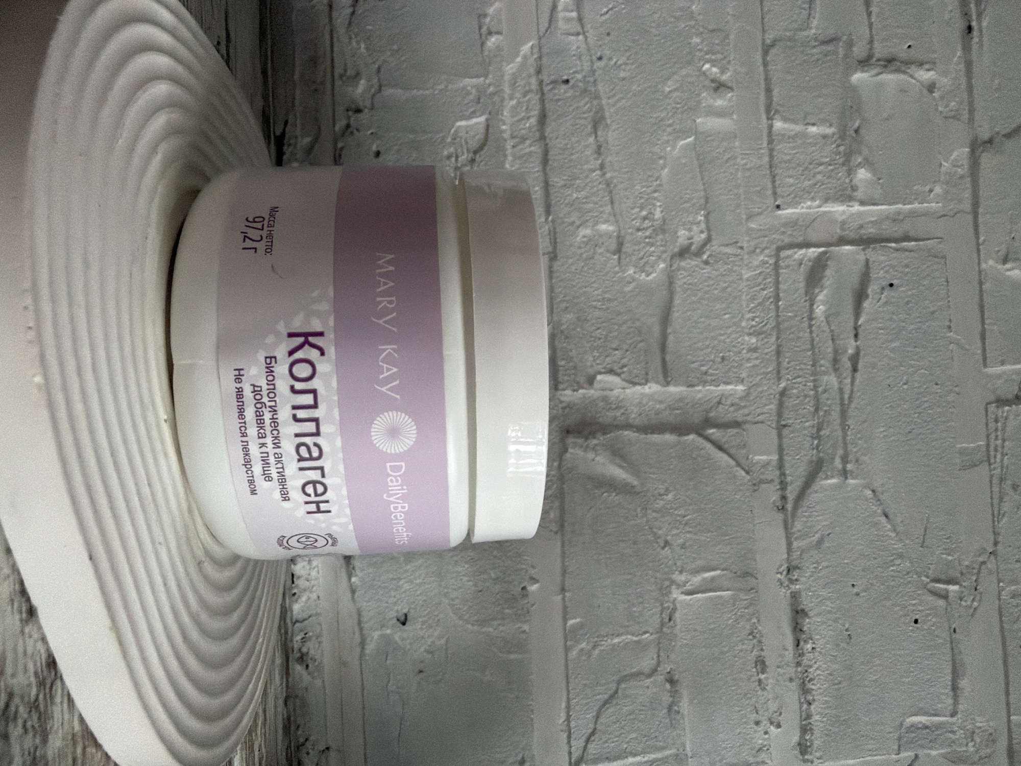 A white, cylindrical jar of Mary Kay collagen supplement is positioned on a white, circular, ribbed surface. The jar has a light purple band around its base with text in Russian and English. The text on the purple band includes "MARY KAY", "DailyBenefits", and "Коллаген" which translates to "Collagen". Below "Коллаген" are the words "Биологически активная" and "добавка к пище" which translate to "Biologically active" and "food supplement" respectively. Further down, "Не является лекарством" translates to "Is not a medicine". On the left side of the jar, the text "Масса нетто:" and "97,2 г" indicates a net weight of 97.2 grams. The background is a white brick wall. The image does not contain any information that would allow for its location to be identified as Dalneretschensk, Russia.