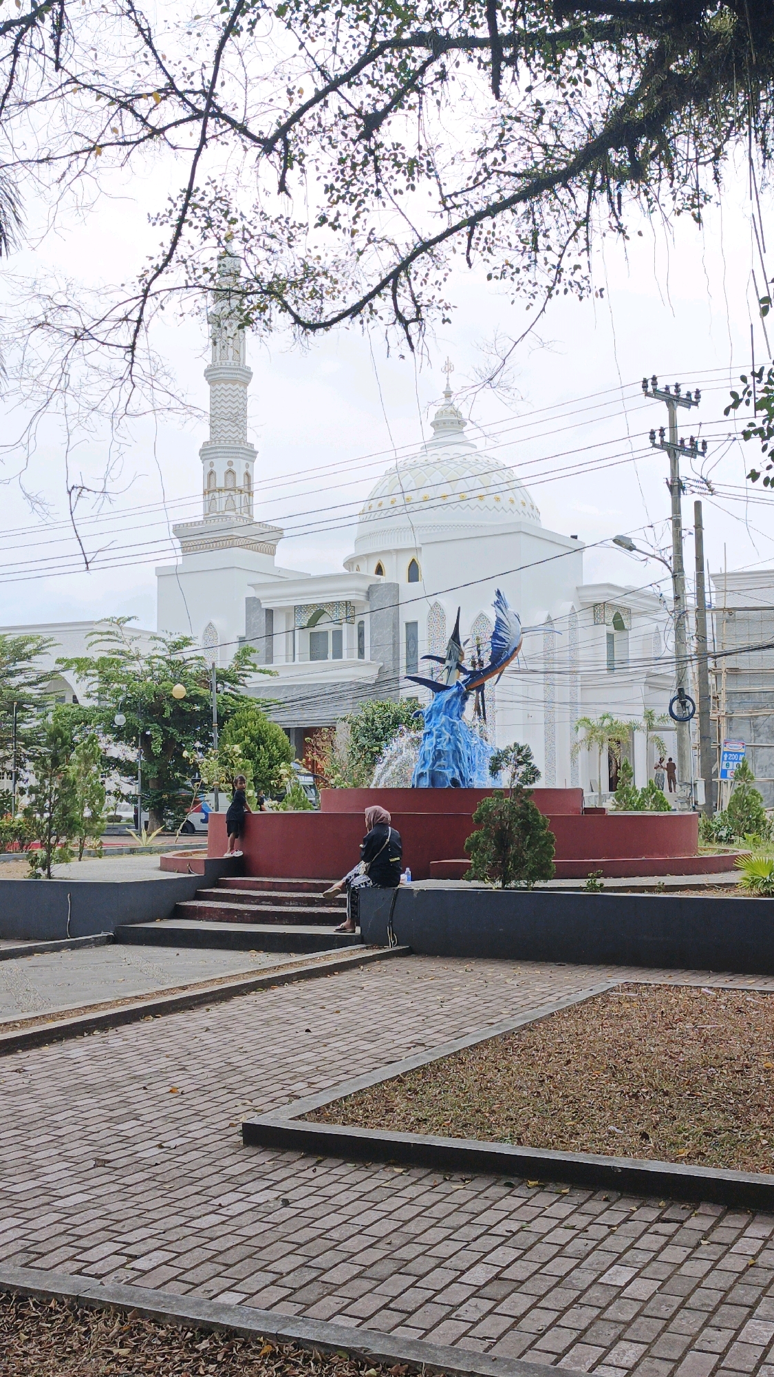 The image shows an outdoor scene in Desa Karangbenda, Indonesia. A white mosque with a large dome and a tall minaret is visible in the background. In the foreground, there is a red circular fountain with a sculpture of two blue winged creatures in mid-air, spraying water. A woman wearing a hijab is sitting on the edge of the fountain's base, facing left. To the left of the fountain, a young child in a black dress stands on the steps leading up to the fountain. The ground in front of the fountain is paved with brick-like stones. Trees and foliage surround the fountain and mosque. Power lines and utility poles are also visible in the background, along with other buildings. The sky is overcast.