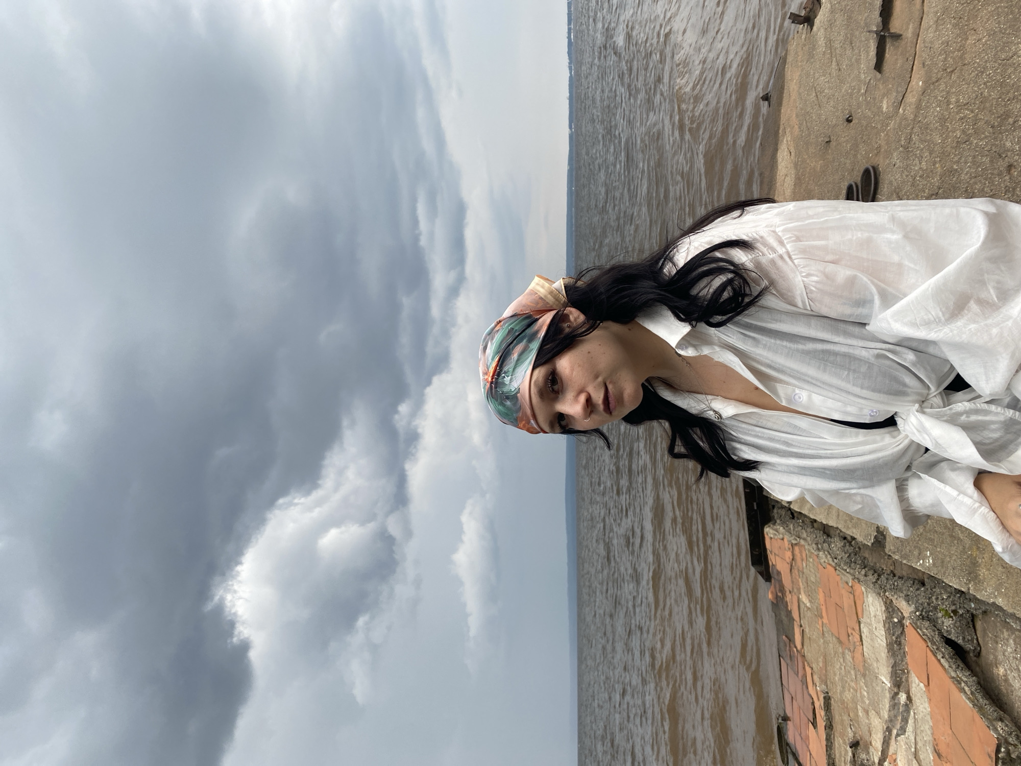 A young woman with dark hair and a colorful headscarf is seated on a concrete structure by the Kama River in Chaikovsky, Russia. She is wearing a white oversized shirt tied at the waist, revealing a black garment underneath. The water is choppy under a cloudy sky, and some weathered bricks are visible on the left side of the frame. The background shows a distant shoreline and some boats. The scene appears to be on a pier or an abandoned industrial area.