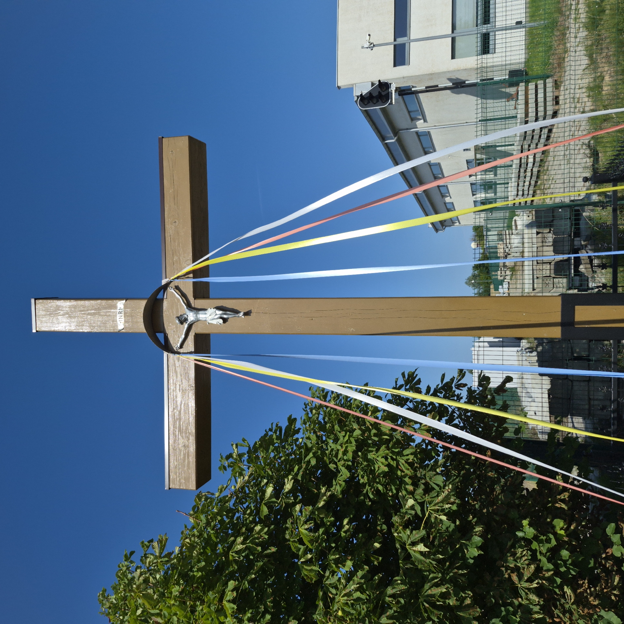 A crucifix stands against a clear blue sky. The crucifix is made of brown wood, with a metal plaque at the top that reads "INRI". A silver figure of Jesus Christ is affixed to the center. Colorful ribbons, including yellow, white, and blue, hang from the cross. To the left, a large green tree with leaves partially obscures the view. In the background, a modern, light-colored building with windows is visible, along with a fence and some construction materials. A traffic light is mounted on a pole next to the building. The scene appears to be outdoors, possibly in a public space.