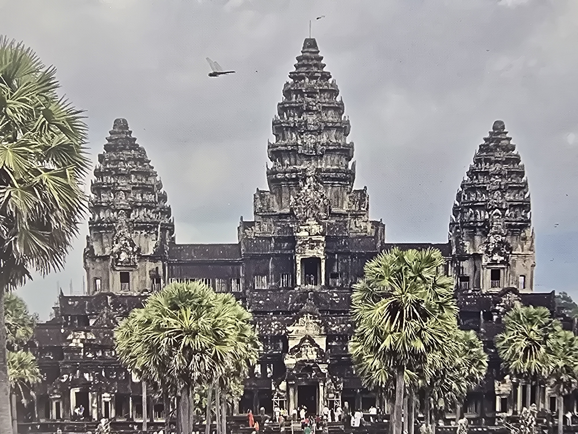 The media file captures a wide view of a large, ancient stone temple complex, clearly identifiable as Angkor Wat in Cambodia. The central subject is the temple, featuring multiple tiered towers (prangs) characteristic of Khmer architecture, with the tallest spire dominating the center. The stone structure appears dark grey and is intricately detailed.
In the foreground and surrounding the temple, several large palm trees with fanned fronds are visible, adding to the tropical setting. Numerous people are gathered at the base of the temple, particularly near the main entrances, suggesting visitors or tourists exploring the site.
The scene is set during the daytime under an overcast, grey sky. A distinctive, dark object with visible wing-like structures, resembling an aircraft or an unidentified flying object, is present in the upper left portion of the sky. No text is visible in the image.
Despite the provided location context of "Sofia, Bulgaria," the architectural style, tropical vegetation, and unique design elements definitively identify the landmark as Angkor Wat, a renowned historical site in Cambodia.