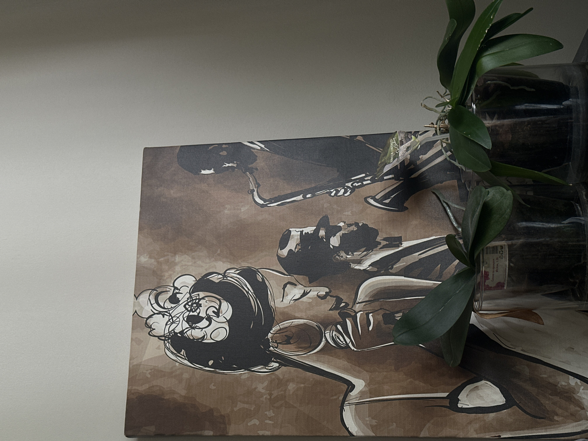 The image displays a canvas painting of a jazz band, featuring a singer and a saxophonist, rendered in shades of brown and black. The painting is positioned on a surface, likely a shelf or table, in front of a plain, light-colored wall, which appears to be indoors. Two clear glass containers holding plants with green leaves are in the foreground, partially obscuring the lower portion of the painting. One of the plant containers has a label attached to it with text that is partially visible, appearing to be in Cyrillic and Latin script. The overall lighting suggests an indoor setting. There are no discernible elements in the image that can be used to identify its location within the city of Sofia, Bulgaria.