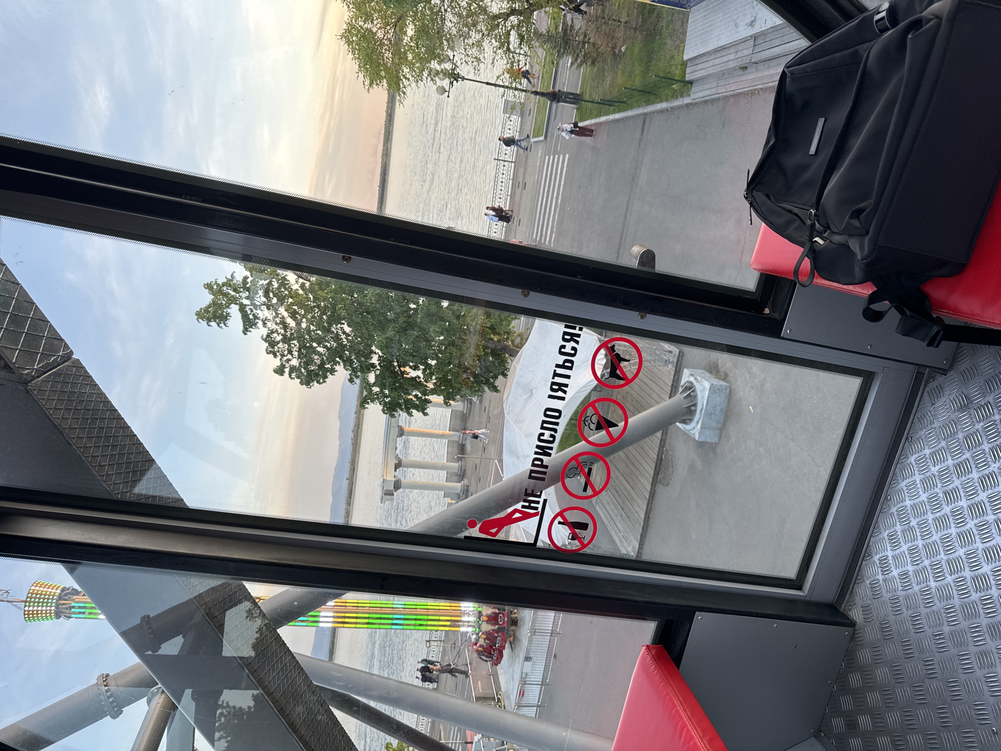 View from inside a Ferris wheel, likely in Khabarovsk, Russia, based on the warning sign written in Russian. A black backpack sits on a red seat. Outside the window, people are walking along a waterfront promenade. A pier with decorative columns extends into the water. The tower and illuminated features of another amusement ride are visible in the reflection of the window. A metal floor is visible inside the Ferris wheel car. The warning sign on the window displays prohibition symbols against climbing, ice cream, smoking, and alcohol.
