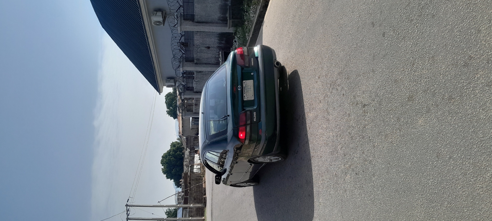 In Jalingo, Nigeria, a dark green 1990s era Mazda 626 sedan is parked on a paved road. The car's brake lights are illuminated. The vehicle has a license plate that reads "GUB-313K" and "110 LAGOS". In the background, there are residential buildings with grey walls and dark corrugated iron roofs, some equipped with air conditioning units and barbed wire fencing. Trees and utility poles are also visible. The sky is clear blue with some wispy clouds.