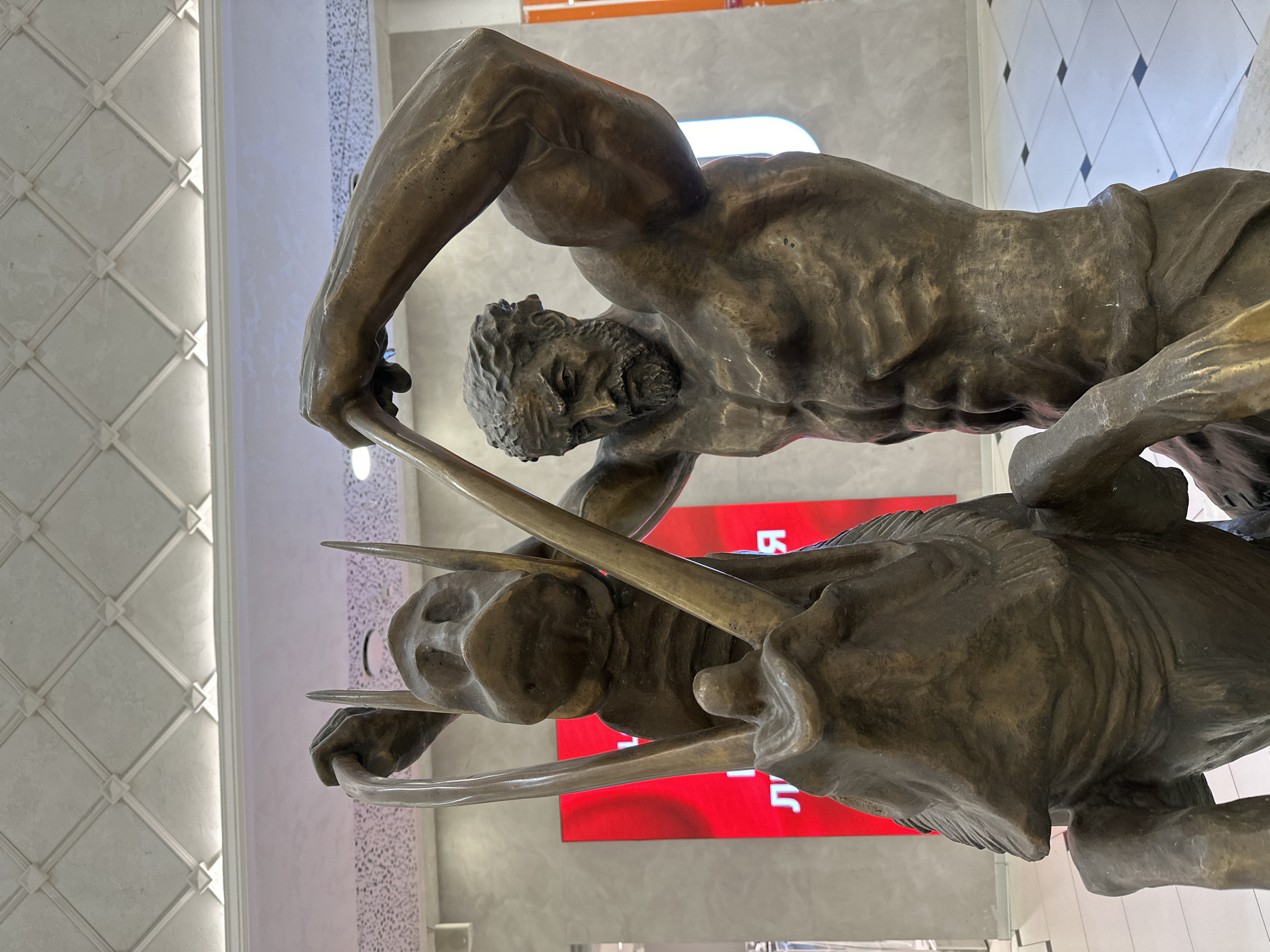The image displays a bronze sculpture of a muscular, bearded man engaged in a dynamic struggle with a large wild boar. The man is depicted in an active pose, wrestling or grappling with the powerful animal, his arms wrapped around its body and his face turned towards it with an expression of exertion and focus. The boar, with prominent tusks, appears to be fighting back or resisting, its form conveying strength and wildness. The sculpture's surface has a rich, textured bronze patina, enhancing the anatomical details of both figures.
The setting is an indoor space, likely a modern public building such as a mall or lobby, in Yekaterinburg, Russia. The background features a pristine white ceiling adorned with a geometric, possibly coffered or paneled, design, illuminated by integrated strip lighting. Behind the statue, a large red display screen is visible, showing fragmented white Cyrillic letters, including "Ь" and "Л." The lighting in the scene is bright and even, highlighting the intricate details of the bronze artwork. The overall impression is one of intense action and heroic conflict, reminiscent of classical mythological themes like Hercules's feats.