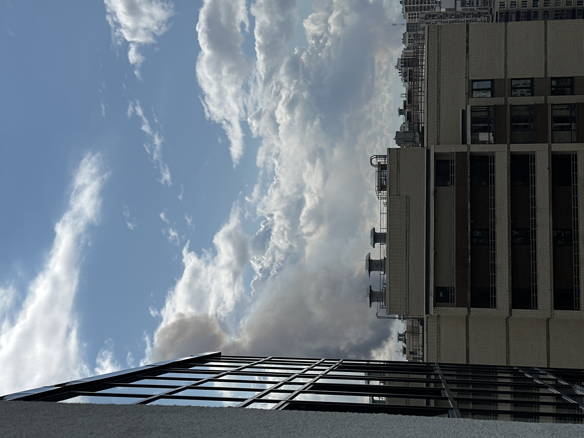 A plume of dark smoke rises into a partly cloudy sky above a cityscape. In the foreground on the left, a modern building with a textured white facade and dark-framed windows dominates the frame. To the right of this building, several multi-story residential buildings with brick exteriors are visible, some featuring balconies. Chimney-like structures are present on the roofs of these buildings. The smoke appears to originate from behind these buildings, obscuring the view of the background cityscape. The sky is a mix of blue and white clouds, with the smoke plume being the most prominent feature against this backdrop. The image appears to be taken from an elevated position, looking out over the city.
