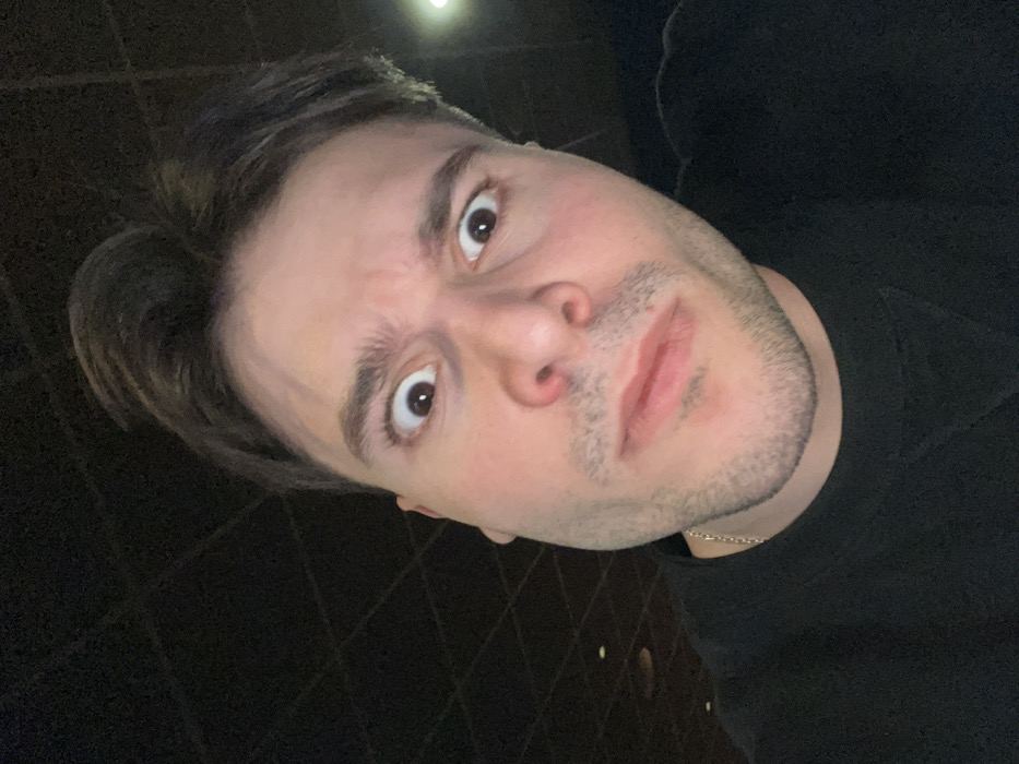 A man gazes upward with wide, expressive brown eyes and slightly furrowed brows. His dark hair is neatly styled, and he has light stubble on his face. He wears a black shirt. A gold chain is visible around his neck. The photo has a slight tilt, making the perspective appear as if taken from below. The background is dark, with a few faint, blurred lights.