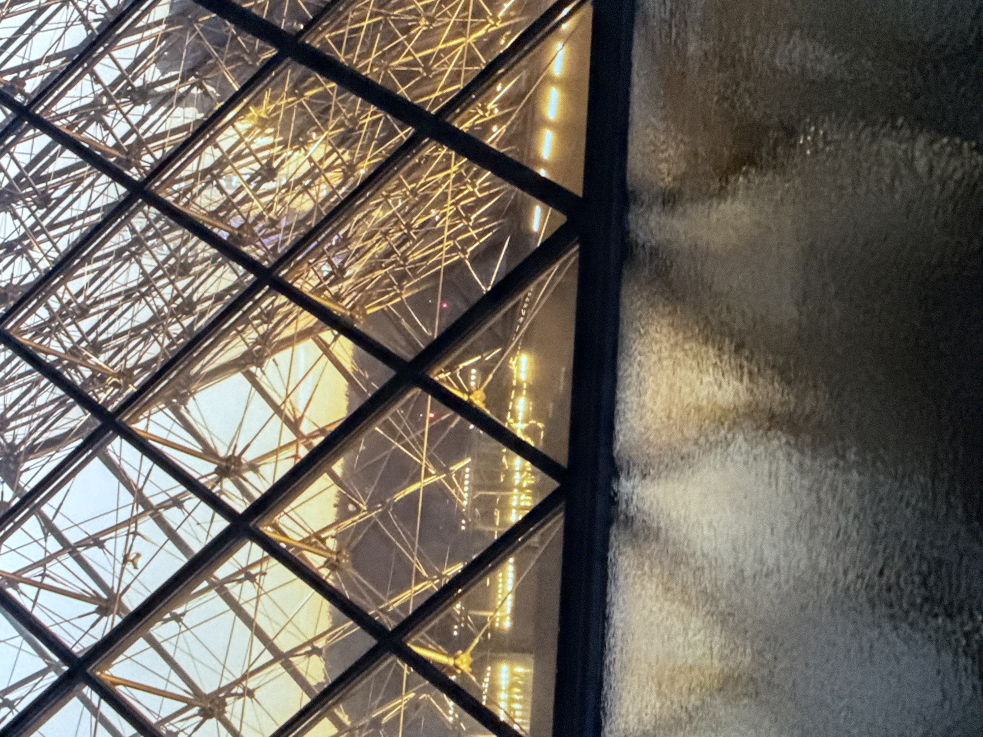 The image captures a portion of the Louvre Pyramid at dusk, as seen from a reflective pool. The geometric structure of the pyramid, composed of glass and metal, is illuminated from within, creating a warm glow that contrasts with the darkening sky. The water in the foreground mirrors the pyramid's lights, distorting them into abstract patterns of light and shadow. The scene is devoid of people and specific geographical markers to place it in Saratov, Russia; the description is limited to the visual elements present in the photograph.