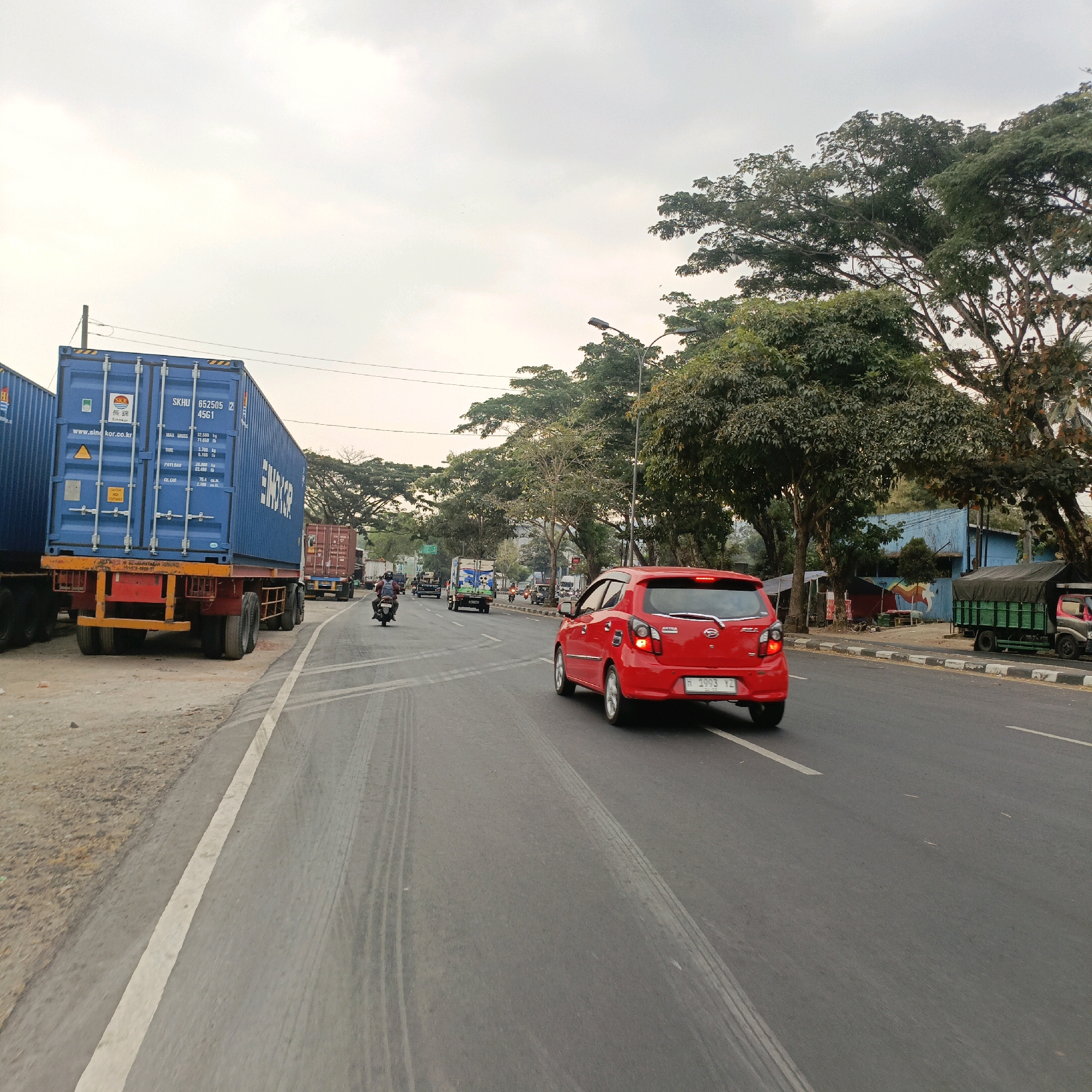 The image depicts a street scene in Salatiga, Indonesia, on an overcast day. In the foreground, a red car is driving on a multi-lane asphalt road, with tire marks visible on the surface. To the left, several large blue shipping containers are parked on a dirt shoulder, adjacent to the road. One of these containers has markings including "SKHU 652505 2 4561" and "SINOKOR.CO.KR". In the background, a variety of vehicles, including trucks and motorcycles, are present on the road. A person is seen riding a motorcycle in the middle ground. Lush green trees line the right side of the road, and a few small buildings are visible behind them. The sky is mostly cloudy, suggesting a potentially humid climate.