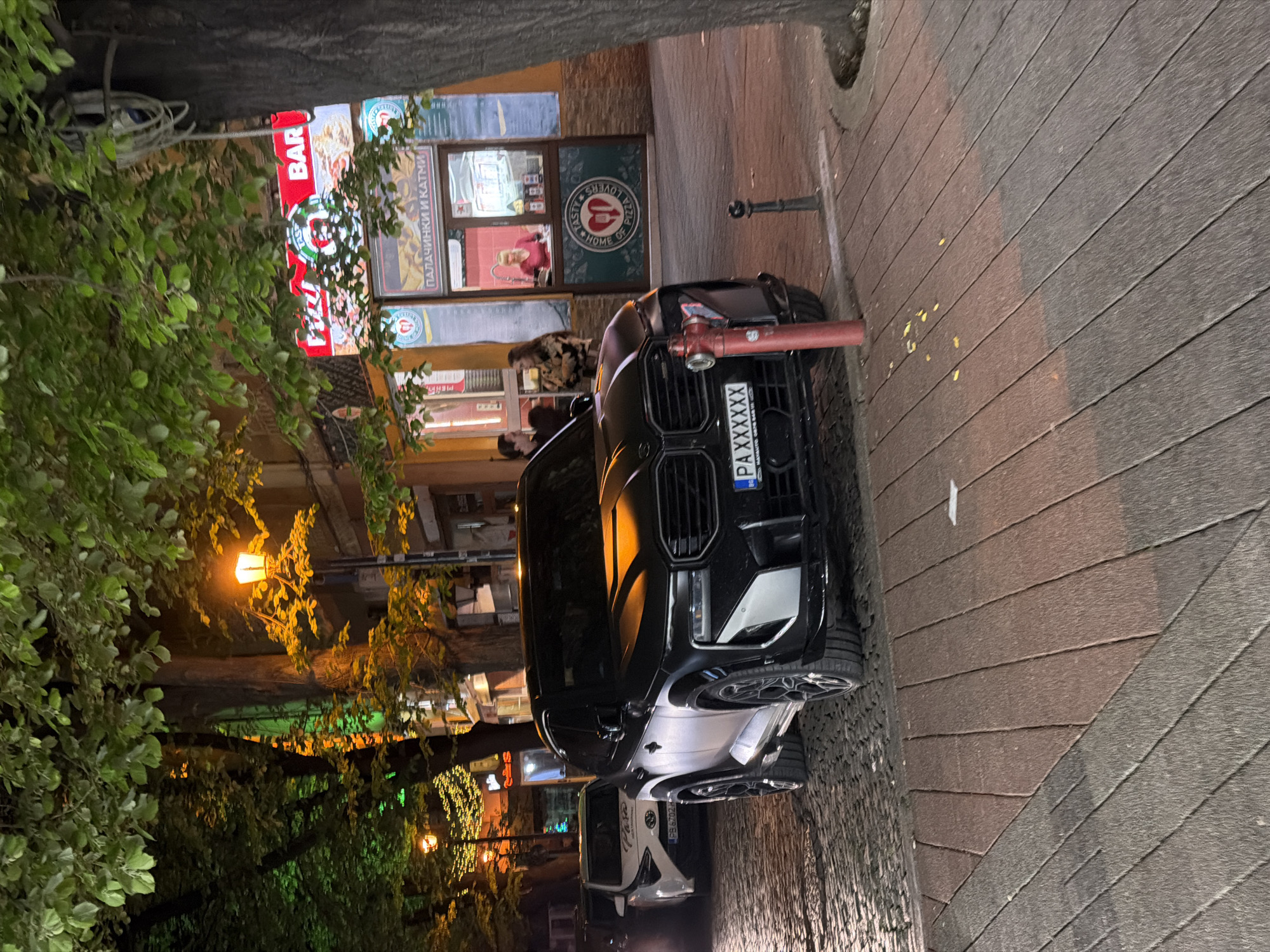 The image depicts a black SUV with a Bulgarian license plate "PA XXXXXX" parked on a cobblestone street in Plovdiv, Bulgaria. The vehicle is partially blocking a red fire hydrant. Behind the car, a "Pizza Lovers" restaurant is visible, with its signage in Bulgarian, including "ПАЛАЧИНКИ И КАТМИ." Patrons can be seen inside the restaurant. To the left, illuminated by streetlights, are trees lining the street. A "BAR" sign is also visible.