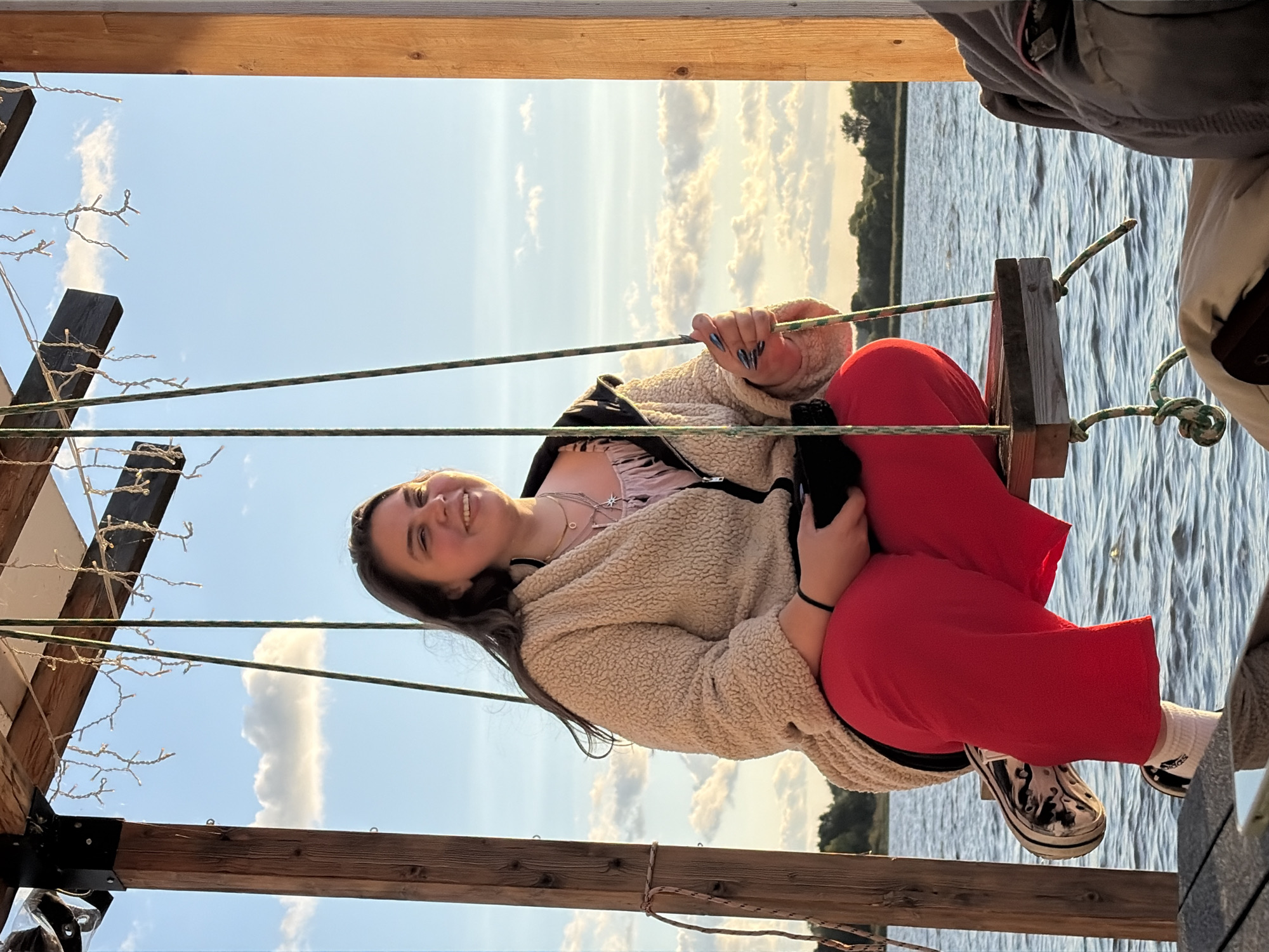 A young woman with long, dark hair is sitting on a swing suspended over water. She is smiling and looking to her right, with her head tilted back slightly. She is wearing a tan, fuzzy jacket over a pink top and bright red pants. Her right hand is resting on her lap, holding a black object, possibly a phone. Her left hand is holding onto the rope of the swing. She is wearing black and white patterned sneakers with white socks.
The swing is attached to a wooden structure with strings of small lights hanging from it. The background shows a body of water, with trees and vegetation visible on the distant shore. The sky is a clear blue with scattered white clouds, and the sun appears to be setting, casting a warm glow over the scene. The scene appears to be an outdoor recreational area, possibly a park or waterfront cafe, in the city of Vidogoshchi, Russia.