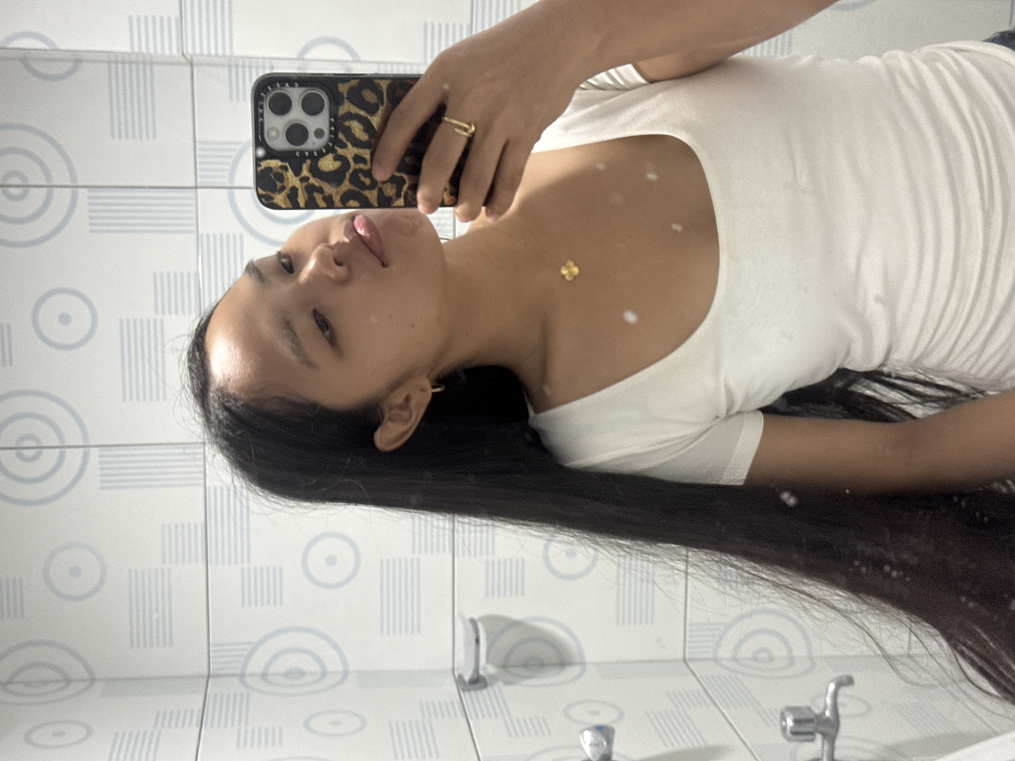 A young woman is taking a mirror selfie in what appears to be a bathroom with tiled walls featuring a geometric pattern of concentric circles and lines. She has long, dark hair and is wearing a white, ruched t-shirt. Around her neck, she has a gold necklace with a clover-shaped pendant, and on her right hand, a gold ring. She is holding a smartphone with a leopard-print case to take the photo. The location of Davao City, Philippines, is not discernible from the image.