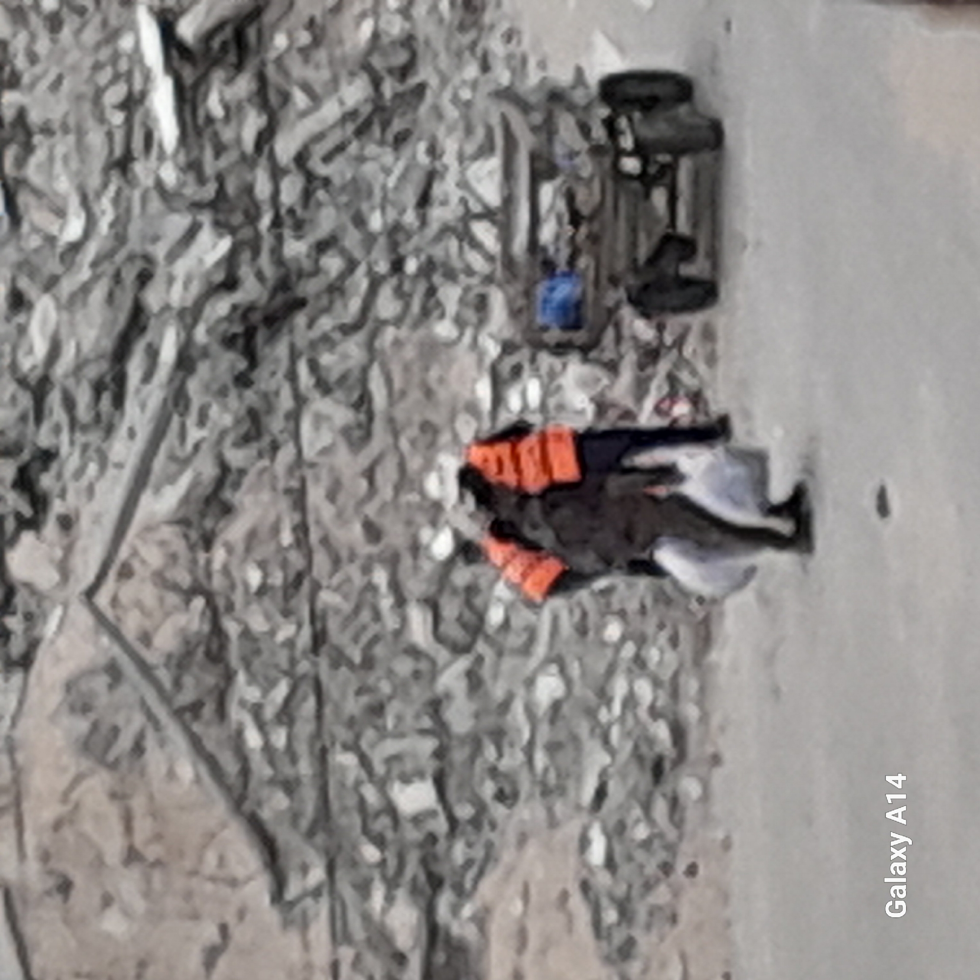 The image shows a scene of devastation in an unknown city. A vehicle is overturned on a gray surface. A person wearing an orange vest lies among rubble and debris. The environment appears to be a construction site or disaster area with scattered gray material.