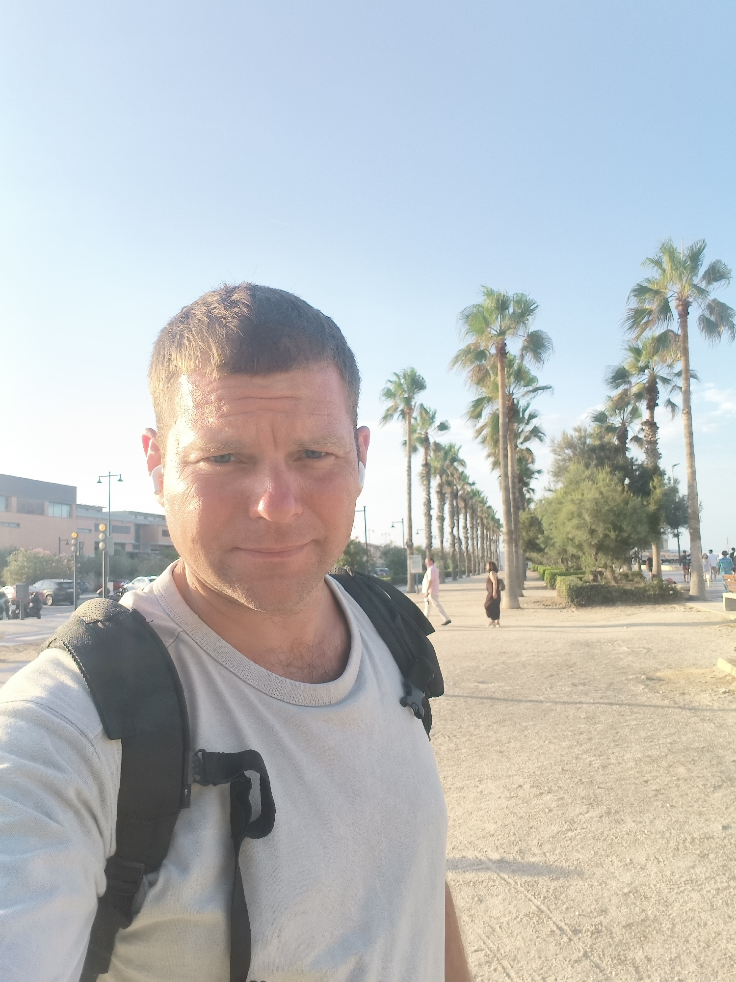 The image is a selfie taken by a light-skinned male with short brown hair and blue eyes, wearing a light grey t-shirt and a black backpack. He has wireless earbuds in both ears. He is standing outdoors on a paved walkway lined with palm trees on the right side and buildings on the left. In the background, several people are walking along the path. The sky is clear and blue. The location is likely the promenade along the beach in the city of Valencia, Spain.