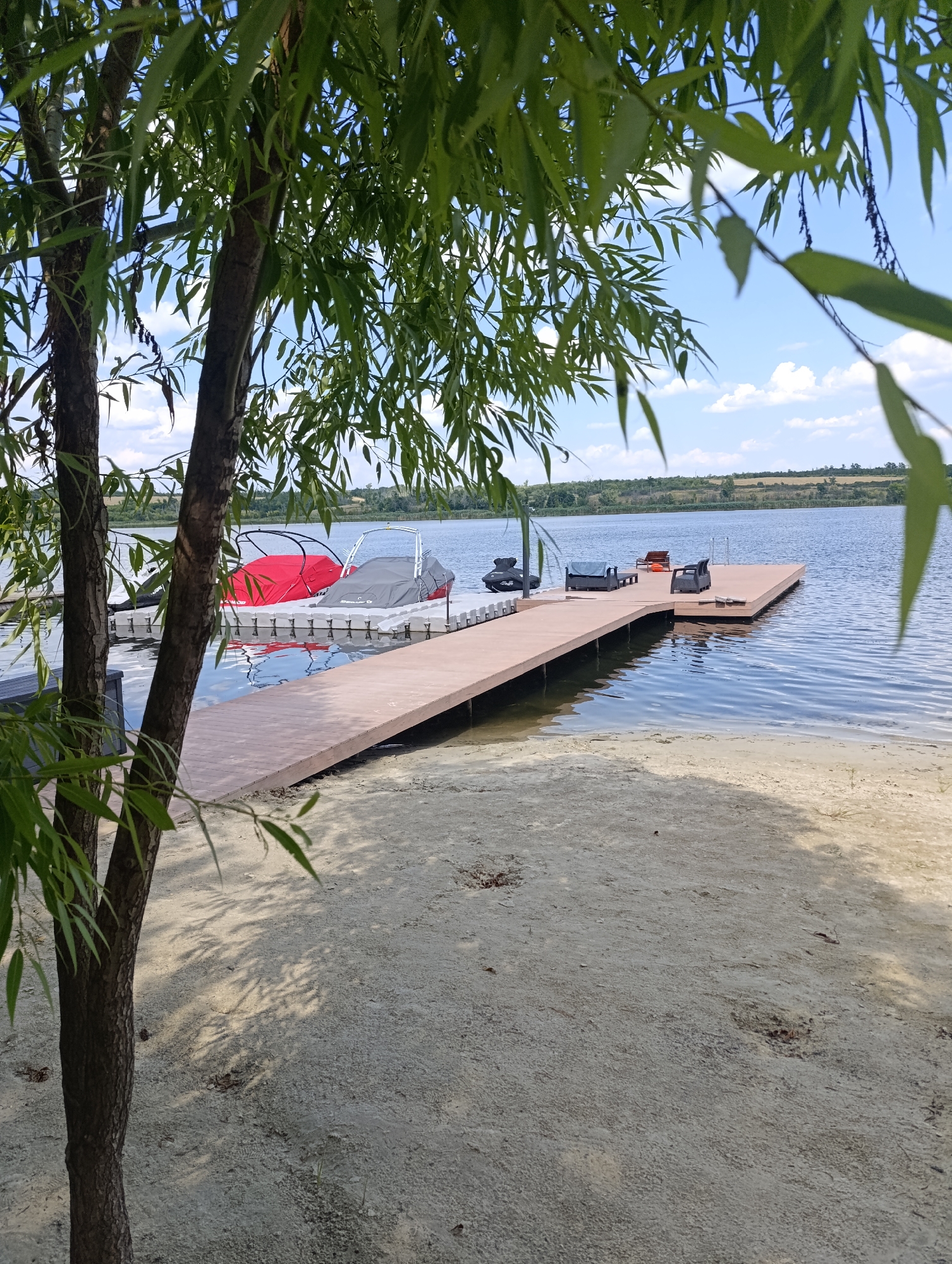 The image depicts a serene lake scene with a wooden dock extending into the water, located on the outskirts of Bulatselivka, Ukraine. The dock is furnished with two black lounge chairs and a small table, suggesting a recreational area. Two boats, one covered in red and the other in gray, are moored to the left of the dock, floating on the calm surface of the lake. Beyond the lake, a gentle slope of green land meets a hazy horizon under a bright blue sky dotted with white clouds. In the foreground, a sandy beach is visible, with the shadow of a tree falling across it. The tree, with its distinctive bark and long, slender leaves, stands to the left of the frame, partially obscuring the view of the boats. The overall impression is one of tranquility and a peaceful natural setting.