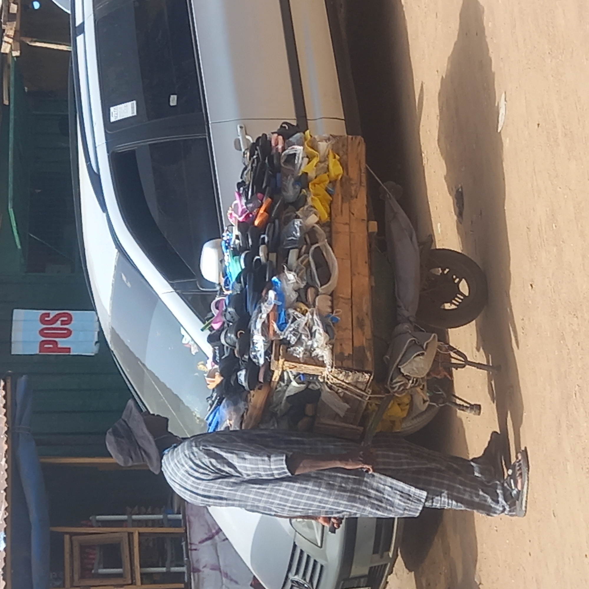 The image, taken in Dadi Riba, Nigeria, portrays an outdoor street or market scene featuring a shoe vendor. In the foreground, a man stands dressed in a dark cap, a light-colored plaid long-sleeved shirt, and matching trousers, with sandals on his feet. He is positioned next to a mobile stall, which appears to be a wooden platform attached to a tricycle or motorbike frame, with a single wheel visible at the back.
This wooden stall is heavily laden with numerous pairs of shoes and sandals of various colors and styles, many of which are individually wrapped in clear plastic bags. The footwear is piled high, indicating a wide range of merchandise. Behind the vendor and his stall, a white car, possibly a sedan, is parked. To the left of the car and partially obscured by it, a green structure features a light blue rectangular sign with the red text "POS" (Point of Sale). The ground is an unpaved, light brown earthen surface. Bright sunlight illuminates the scene, casting distinct shadows, suggesting it is daytime with clear weather. The overall setting is consistent with a vibrant street market environment.