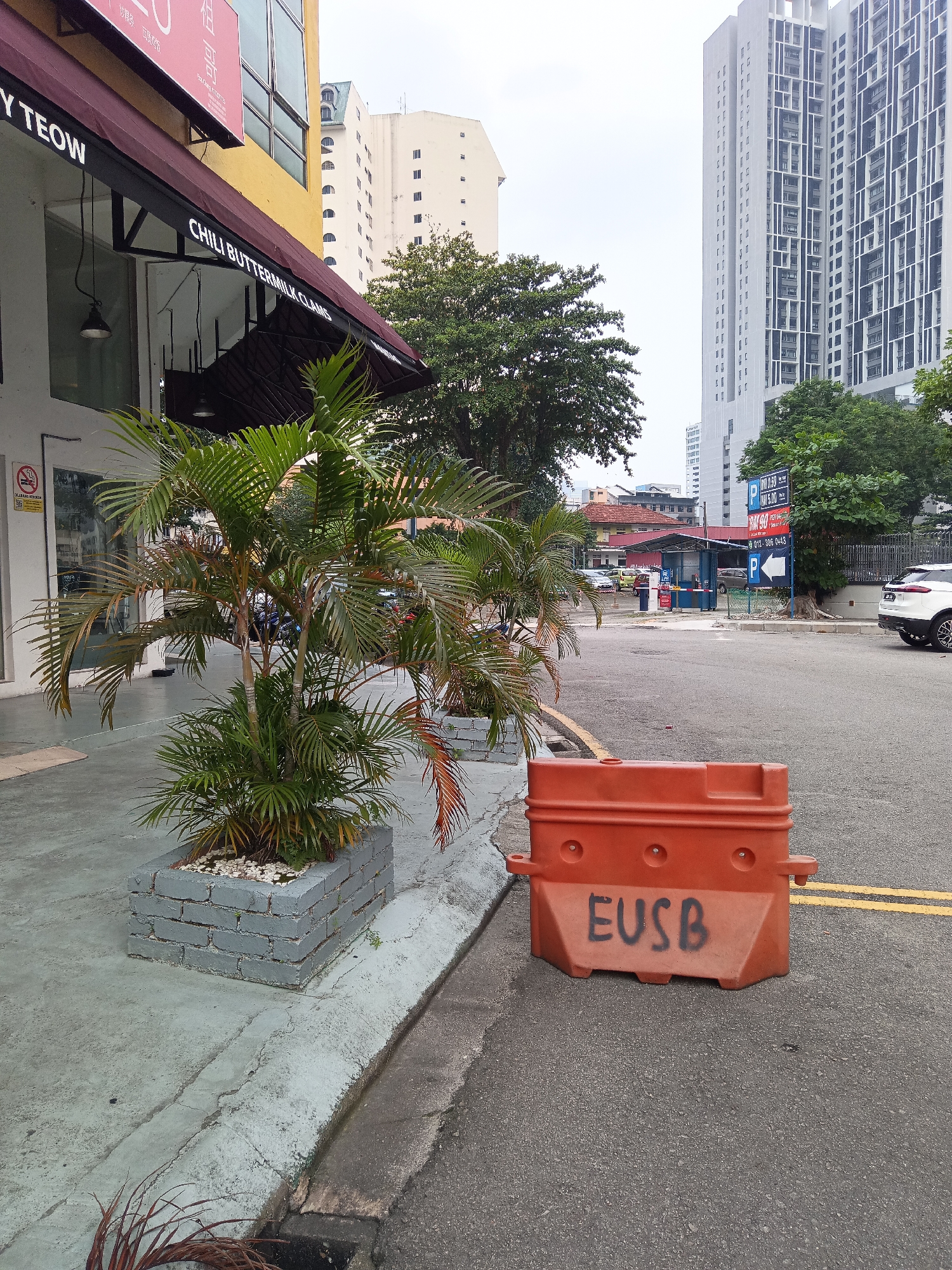 The image depicts a street scene in Kuala Lumpur, Malaysia. In the foreground, an orange traffic barrier with the graffiti "EUSB" is visible on an asphalt road marked with yellow lines. To the left, a concrete sidewalk is present, featuring a planter box containing a palm tree. Adjacent to the sidewalk is a building with a maroon awning displaying text, including "CHILI BUTTERMILK CLAMS". Further in the background, several buildings are visible, including a tall, modern skyscraper and a lower, older structure with red roofing. Trees line the street, and a parking booth with barrier gates is also present. A white SUV is parked on the right side of the road. The sky is overcast.