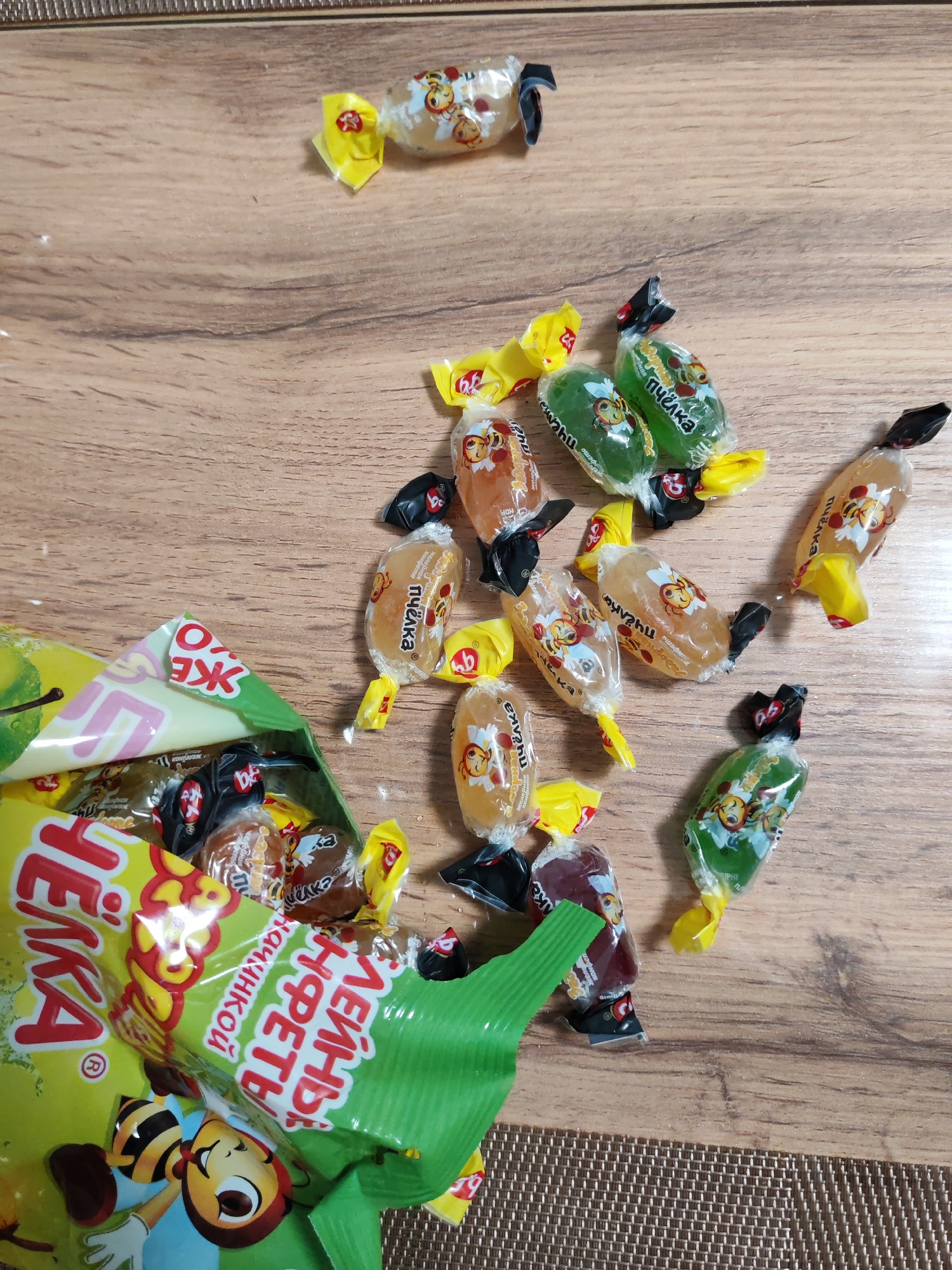 The image shows a pile of individually wrapped candies scattered on a wooden surface, likely a table or floor. The candies are of various colors and flavors, including green, orange, and brown. Some of the candies are still in their original packaging, while others have been opened. A partially opened bag of candies, also green, is visible on the left side of the image. The bag features a cartoon bee character and Russian text, suggesting it is a product sold in Russia. The overall scene depicts a casual and slightly messy arrangement of sweets. The image does not provide any information about the location in Podolsk, Russia, or any people.