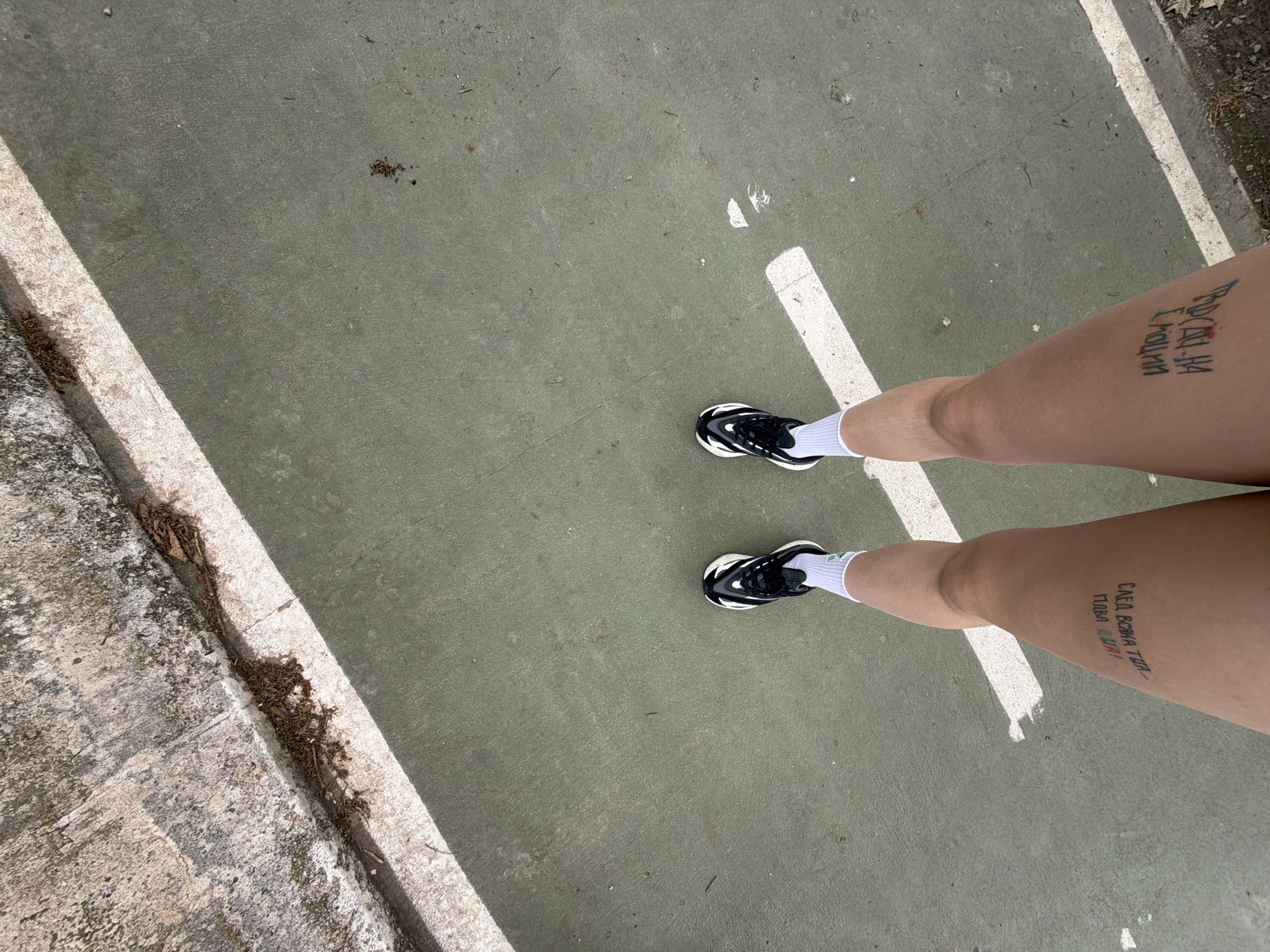 A person stands on a light green painted surface, possibly a sports court, marked with white lines. The location is Arroyo de la Miel-Benalmádena Costa, Spain, as indicated by metadata. The person wears white socks and black and white sneakers. Tattoos are visible on the person's thighs, featuring text in Cyrillic script. The ground surrounding the court has a concrete or stone border with debris present. The scene appears to be outdoors in daylight.