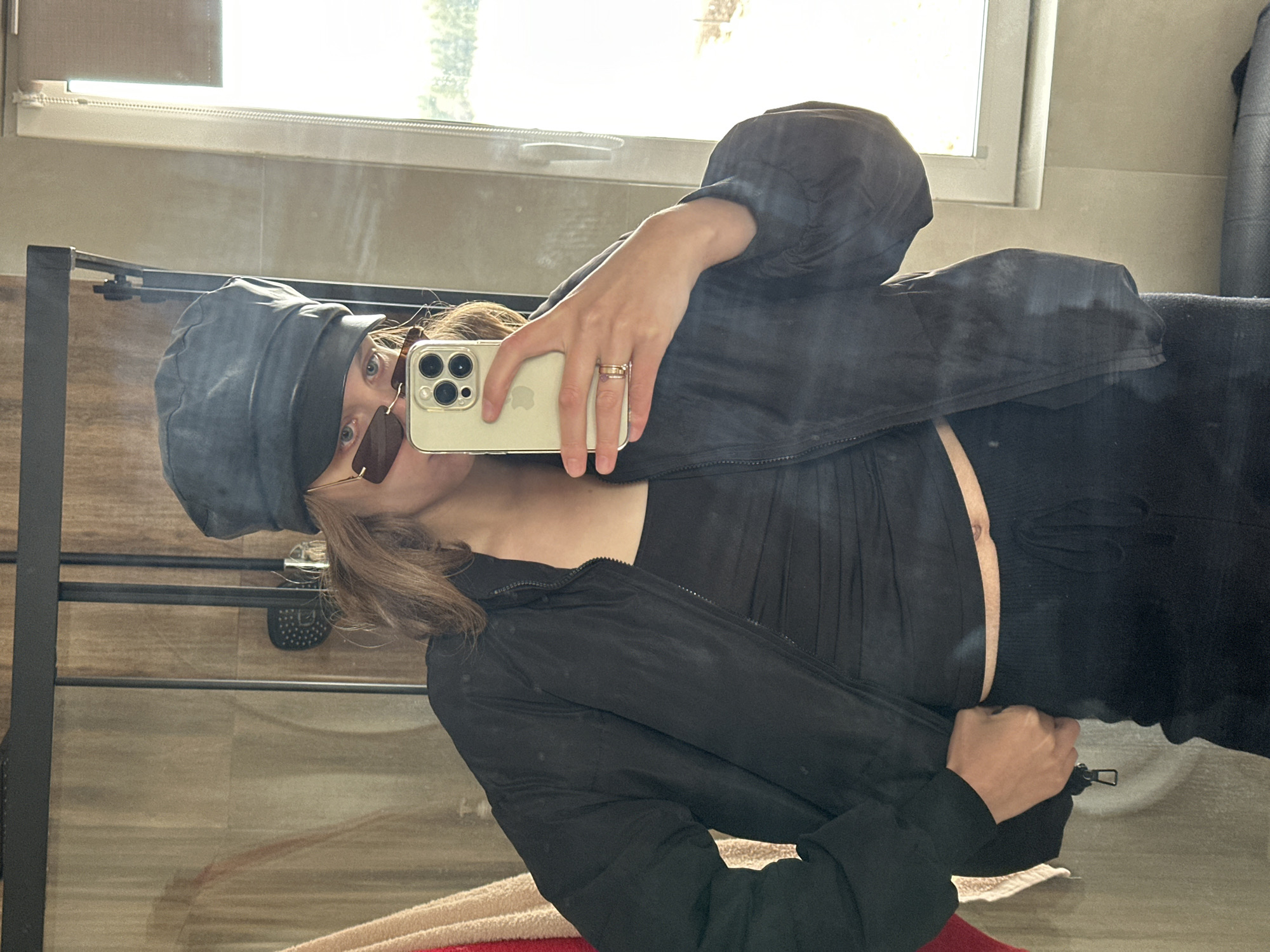 A person in a black outfit, including a cap and jacket, takes a selfie in a bathroom. The individual holds a gold iPhone with multiple rings on their fingers. Reflections in the mirror reveal the bathroom setting, featuring a window and a towel rack. The location is Unknown, Unknown.