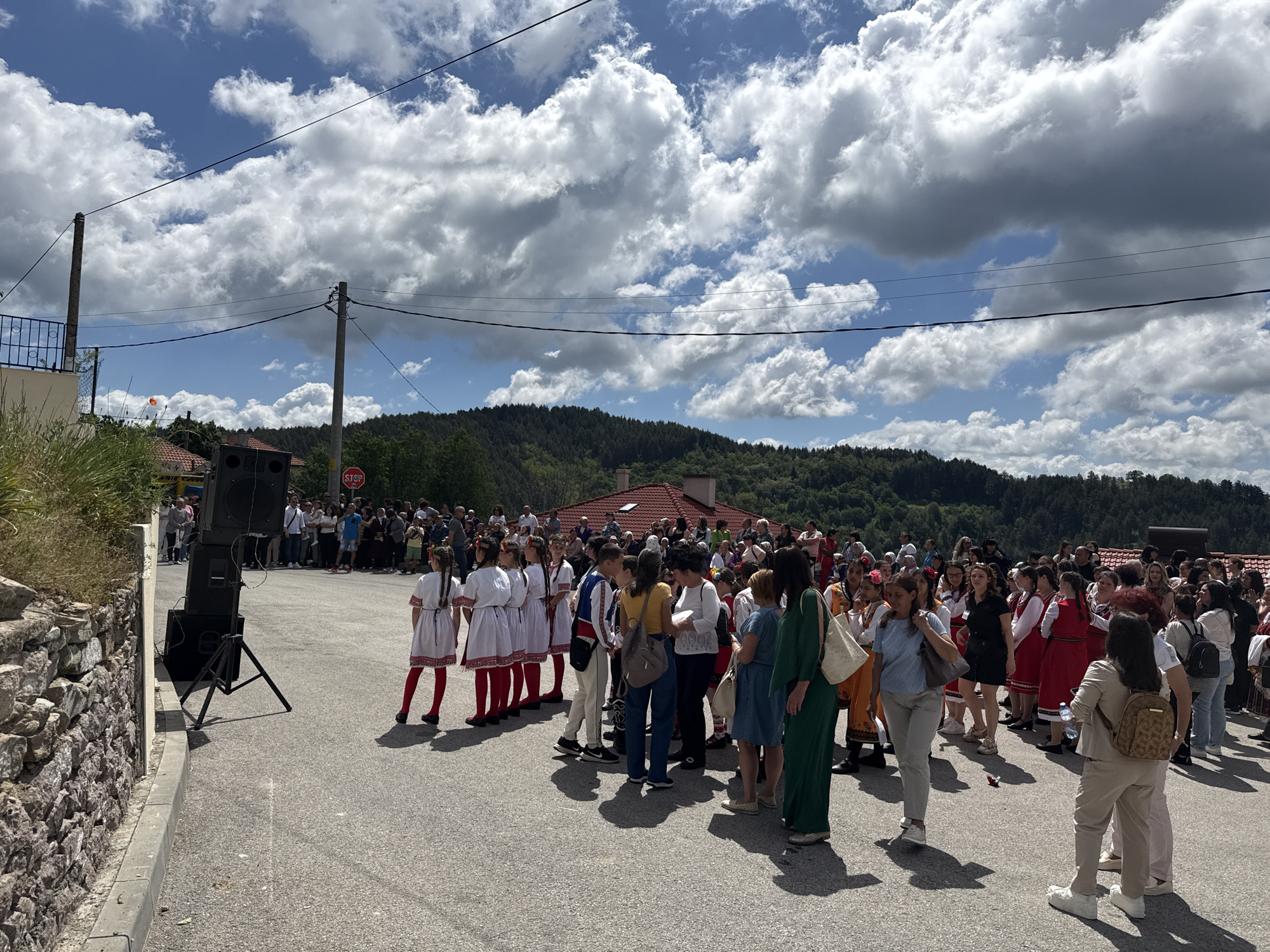 The image shows a gathering of people in what appears to be a village square or a large open area in Pripek, Bulgaria. A substantial crowd of people are present, many dressed in traditional Bulgarian clothing, specifically white dresses with red embroidery and leggings.
A loudspeaker system stands to the left side of the frame, suggesting the event may involve music or announcements. A stop sign is visible in the background, offering a clue about the location being a crossroads. The setting is sunny, with a partly cloudy sky above. The landscape includes a mix of buildings with red tile roofs and forested hills in the background. The ground is asphalt and appears to be a parking or gathering space.