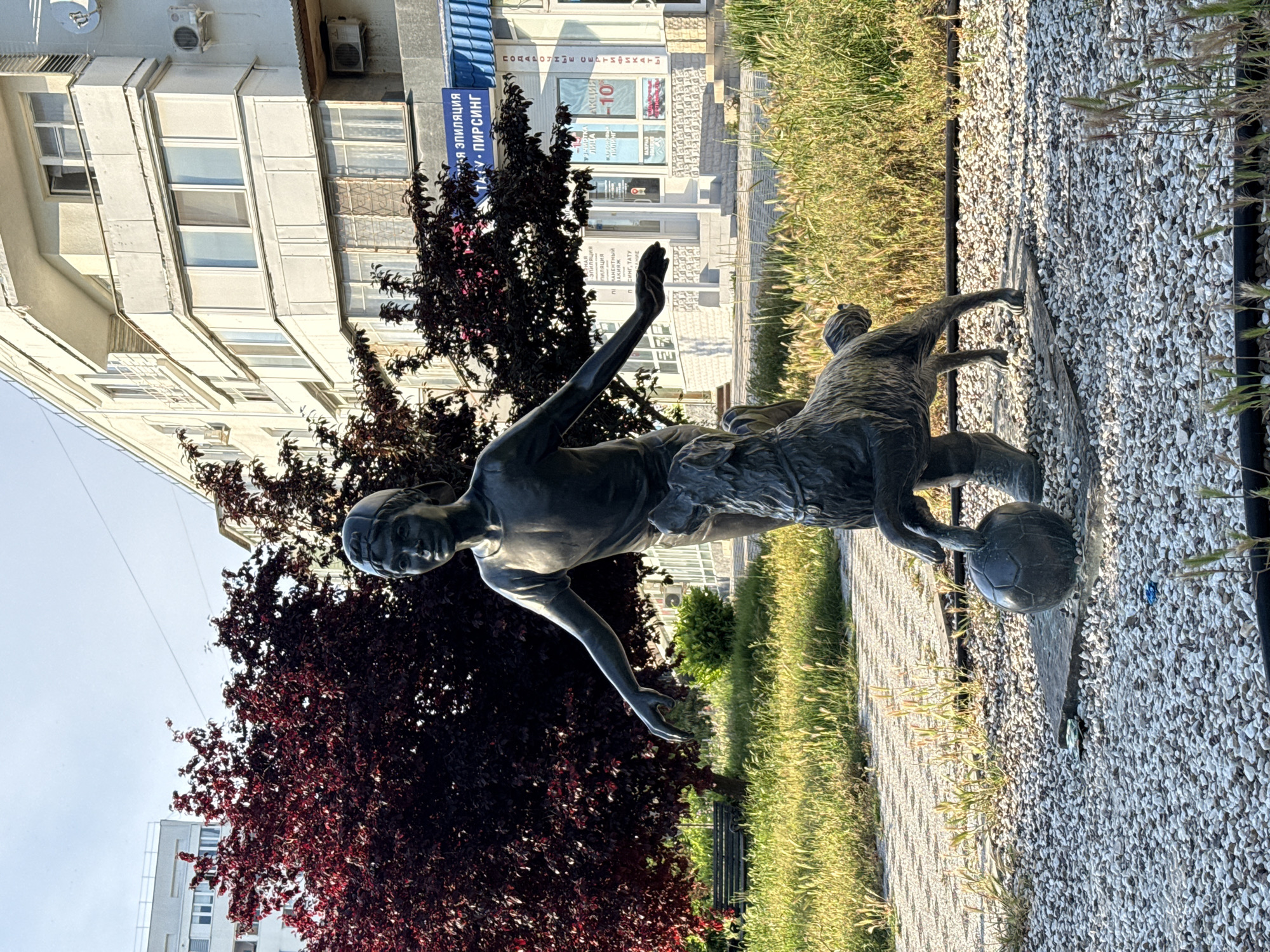 A bronze statue of a boy in a cap and a dog are in the foreground of the image. The boy's right arm is outstretched, and his left hand is on the dog's head. The dog is standing on its hind legs, with its front paws on a soccer ball. The statue is in a park, with a gravel path and green grass. In the background, there is a building with several windows and balconies. The sky is clear and blue. The scene is illuminated by the sun.