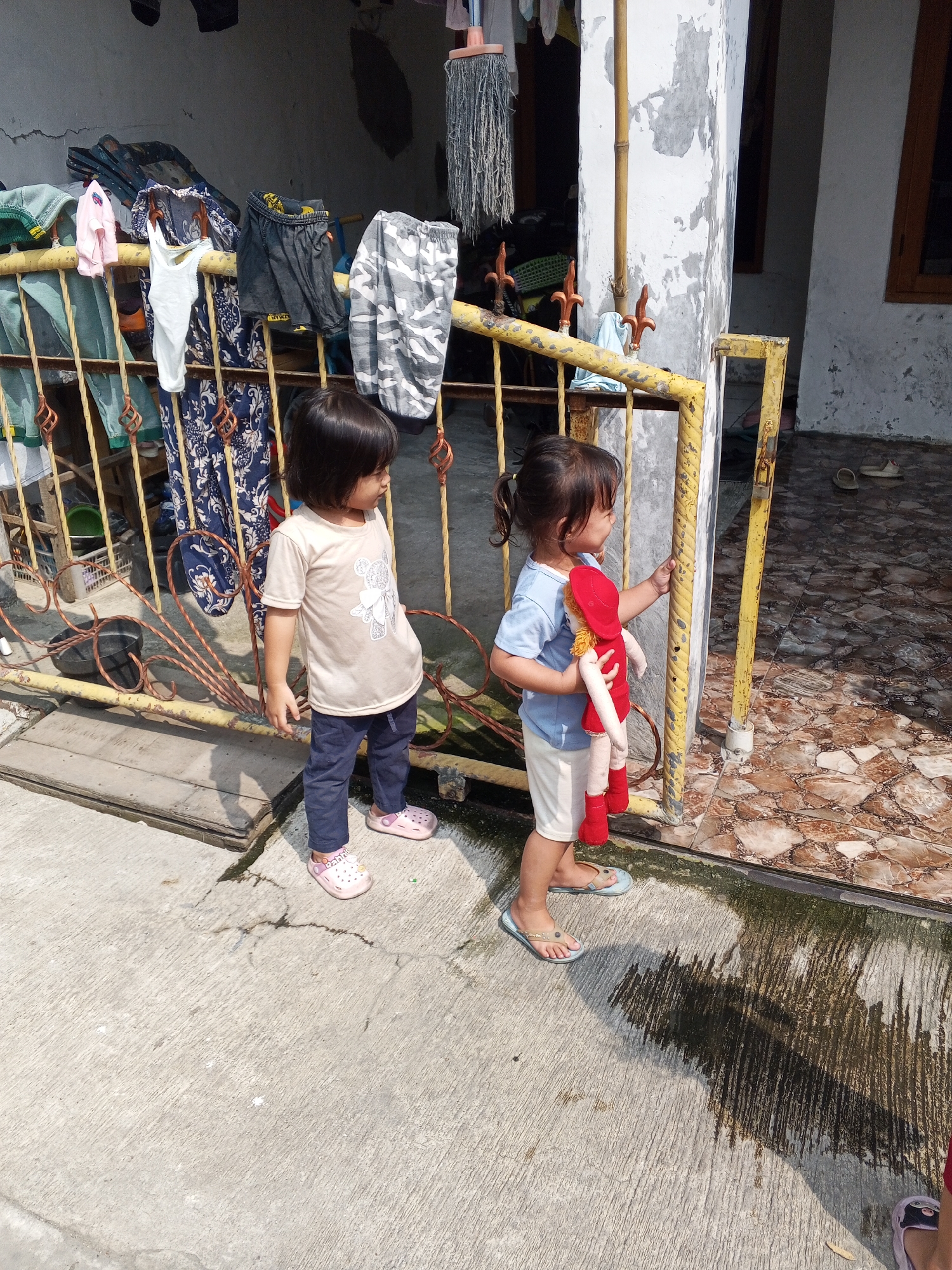 Two young children stand on a concrete walkway in front of a yellow gate. The child on the left, facing forward and slightly to the right, wears a tan t-shirt with a graphic and dark pants, and pink clogs. The child on the right, facing left and holding a red doll, wears a blue t-shirt, white shorts, and light blue sandals. Behind the children, laundry hangs on a clothesline strung between a building and a metal railing. The ground is a mix of concrete and patterned tile. The overall setting appears to be an outdoor residential area.
