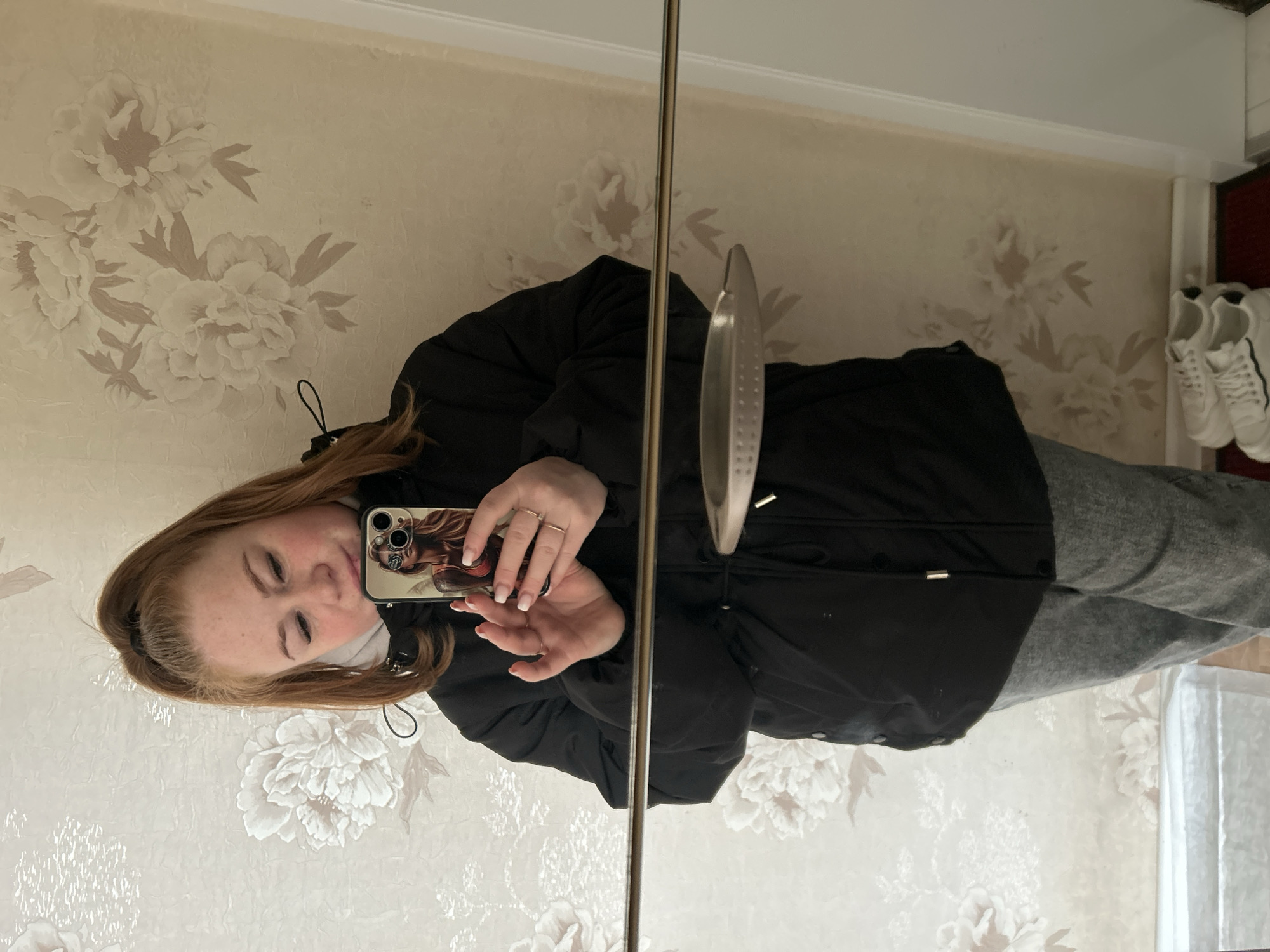 The image shows a woman taking a selfie in a mirror. She is wearing a black jacket and gray pants. She is holding a smartphone with a decorative case. The background is a wall with a floral pattern. There are white sneakers visible in the background, near the corner of the wall and the doorway. The location is likely inside a room.
Given the prompt, it is not possible to determine if the location is Chaikovsky, Russia.