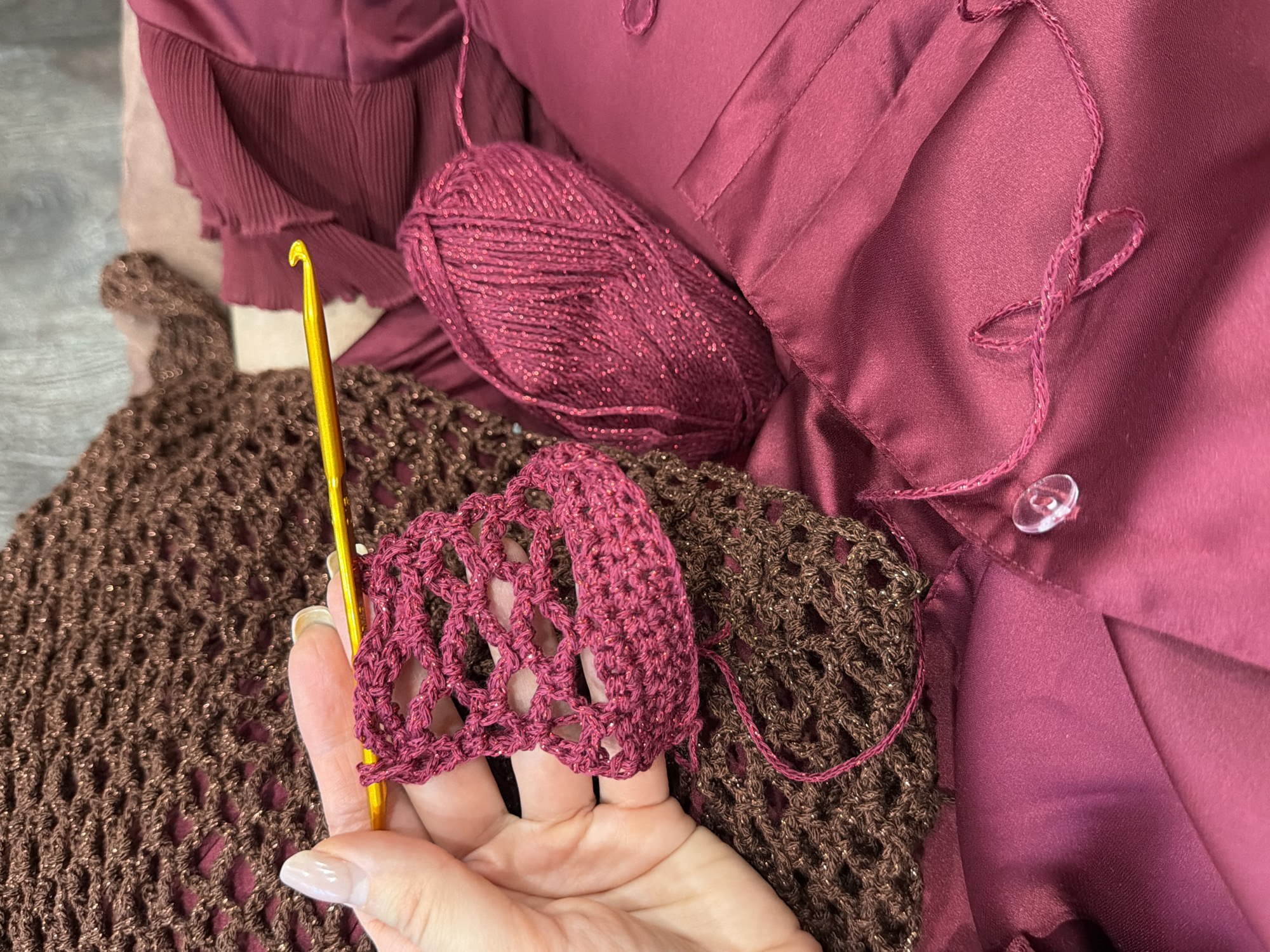 A close-up shot shows a person's hand holding a partially crocheted item, a small section of magenta-colored yarn with a metallic thread, a gold-colored crochet hook, and a larger crocheted piece in brown with a metallic thread. The yarns are presumably the same kind, but different colors. The person is wearing a burgundy-colored top or garment, some of which has a pleated texture. A clear button is visible on the garment. A ball of magenta yarn also rests on the burgundy fabric. The environment appears to be indoors, with the focus on the crafting activity. There is no evidence to suggest that this photo was taken in the city of Krasnoyarsk, Russia.