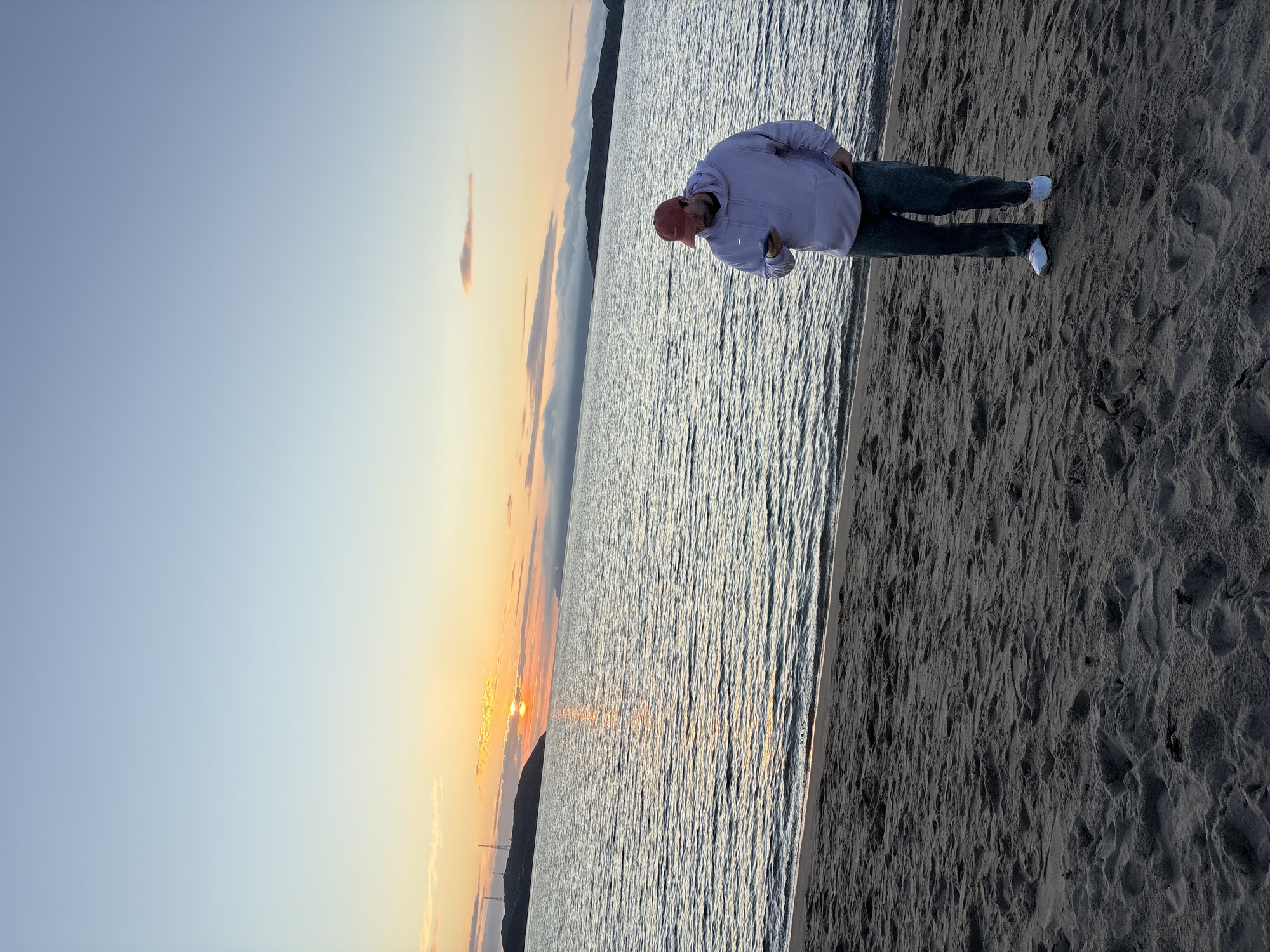 A lone man, wearing a red baseball cap, a light purple hoodie, and dark pants, stands on a sandy beach, looking down at his phone. The ocean stretches out behind him, with gentle waves lapping at the shore. In the distance, a body of land with hills and structures is visible, silhouetted against a sunset sky. The sky is a gradient of soft blues and oranges, with wispy clouds scattered across the horizon. The scene appears to be a tranquil evening on the coast, possibly in the city of Teriberka, Russia, given the characteristic Arctic landscape and the quality of light during a sunset.