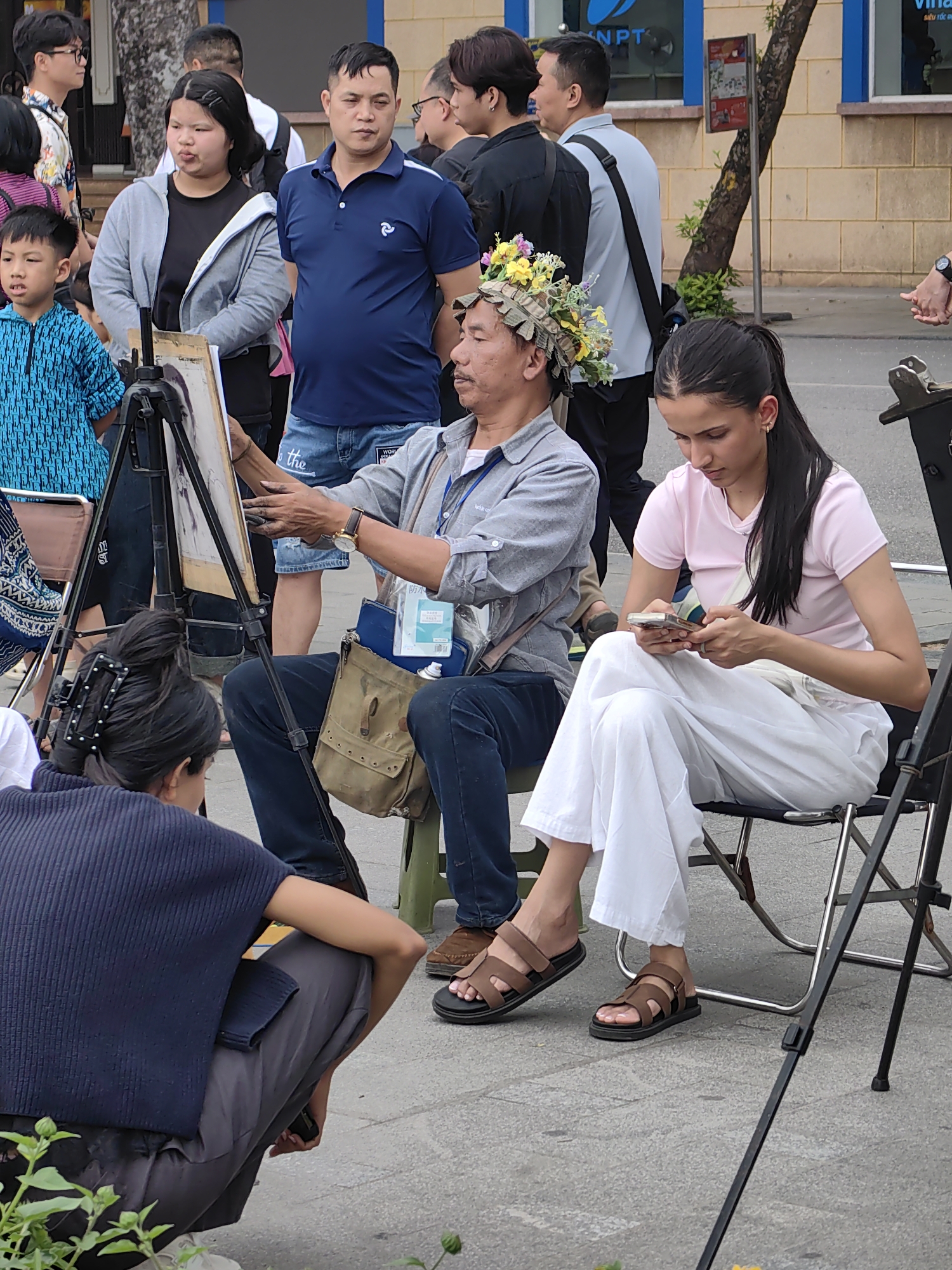 Captured in Hà Nội, Vietnam, the scene features an outdoor art market. An artist, donned with a flower-adorned hat, sits facing an easel, holding a pencil in one hand and an artboard in the other, presumably creating a portrait. Seated next to him is a young woman with dark hair in a ponytail, dressed in white pants and a light pink t-shirt, engrossed in her phone. In front of the artist, a person wearing a dark blue top and pants squats, observing the artist at work.
A diverse crowd stands in the background, including a woman in a gray hoodie, a man in a blue polo shirt, and several others casually dressed. A young boy in a blue patterned shirt stands beside them. The scene is set against a backdrop of a building with windows and a tree.
Additional details include a brown satchel bag next to the artist, sandals worn by both the artist and the woman using her phone, and art supplies scattered around the artist. A logo can be seen on the building in the background.