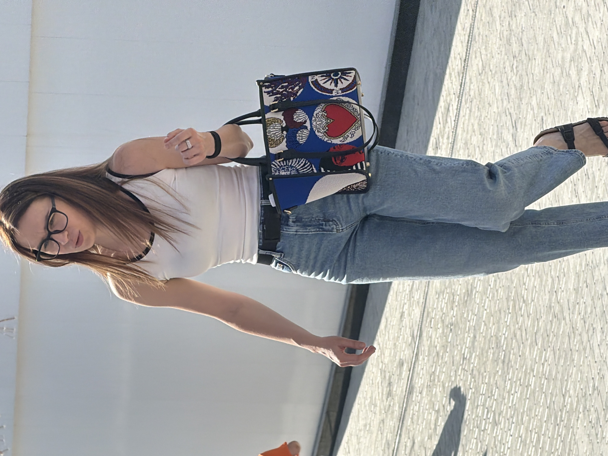 A woman with long brown hair and glasses is walking outdoors, facing slightly to the left. She is wearing a white tank top with black trim, light blue jeans, a black belt, and black sandals. She is carrying a patterned tote bag with blue, red, white, and black designs, featuring a red heart. A ring is visible on her left hand. The background consists of a white wall and a paved ground with a shadow of a person on the left. The scene appears to be in an urban environment.