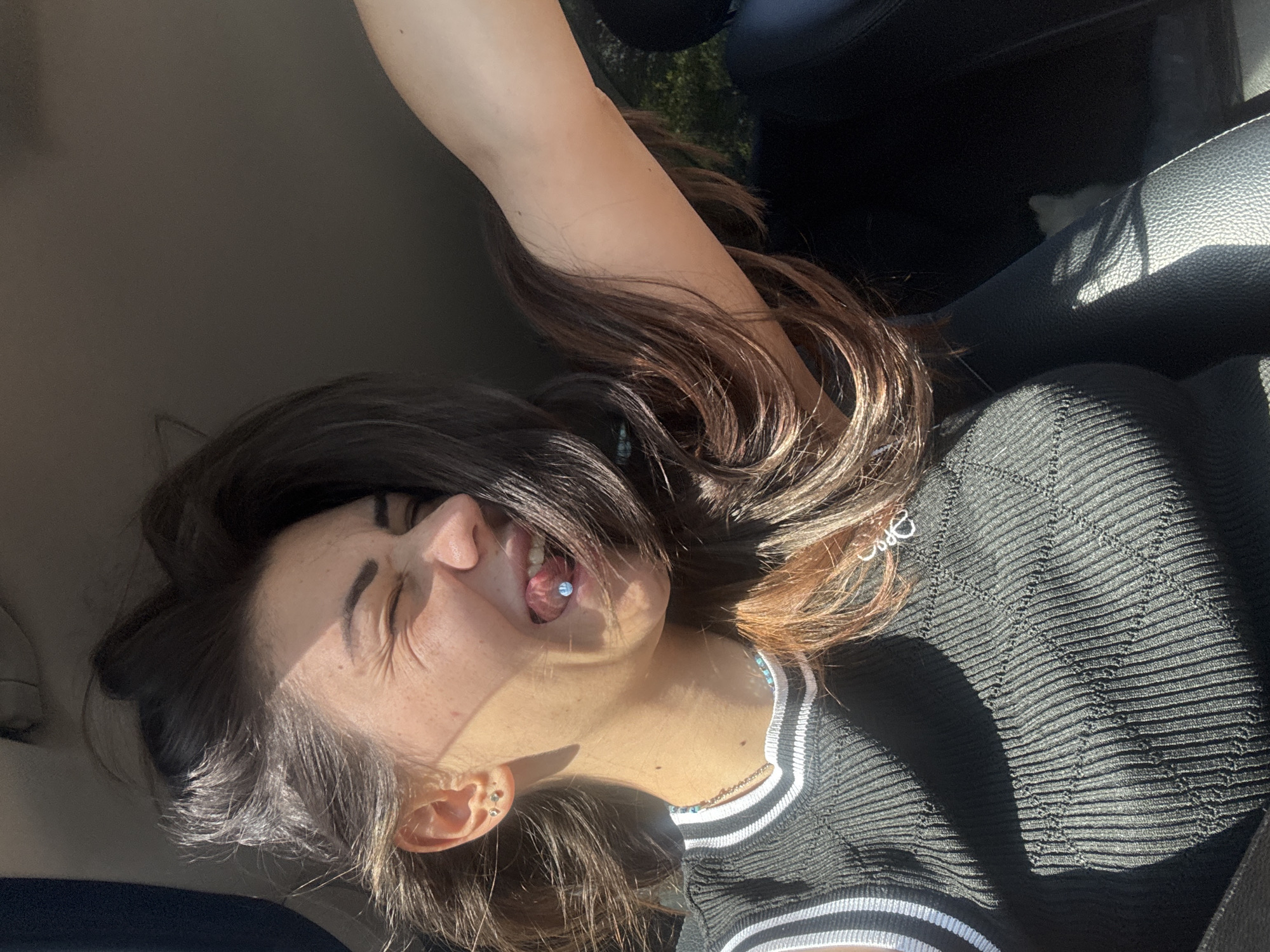 A young woman with long, dark brown hair sticks her tongue out, revealing a blue tongue piercing. She is sitting in the passenger seat of a vehicle, possibly in Sofia, Bulgaria, as indicated by the presence of what appears to be a black car interior with a leather seat. Sunlight streams in, illuminating her face and casting shadows on her black, ribbed top. Her eyes are closed, and she is smiling, suggesting she is enjoying the ride or the moment. Her arm is extended, possibly resting on the car door or window.
