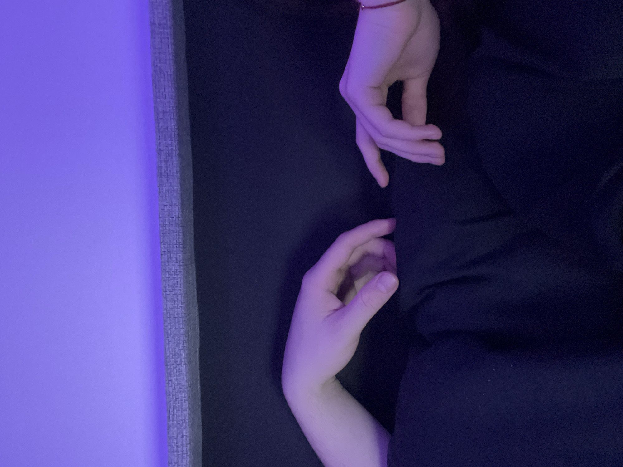 Two hands are visible against a dark blue or black fabric background. The hands appear pale, illuminated by a purple light source coming from the left of the frame. One hand is positioned above the other. The backdrop appears to be fabric with draped folds. A section of what appears to be a grey wall or textured surface is visible on the left edge of the frame, also illuminated by the purple light. There is no information available to suggest the photo was taken in Saint Petersburg, Russia.