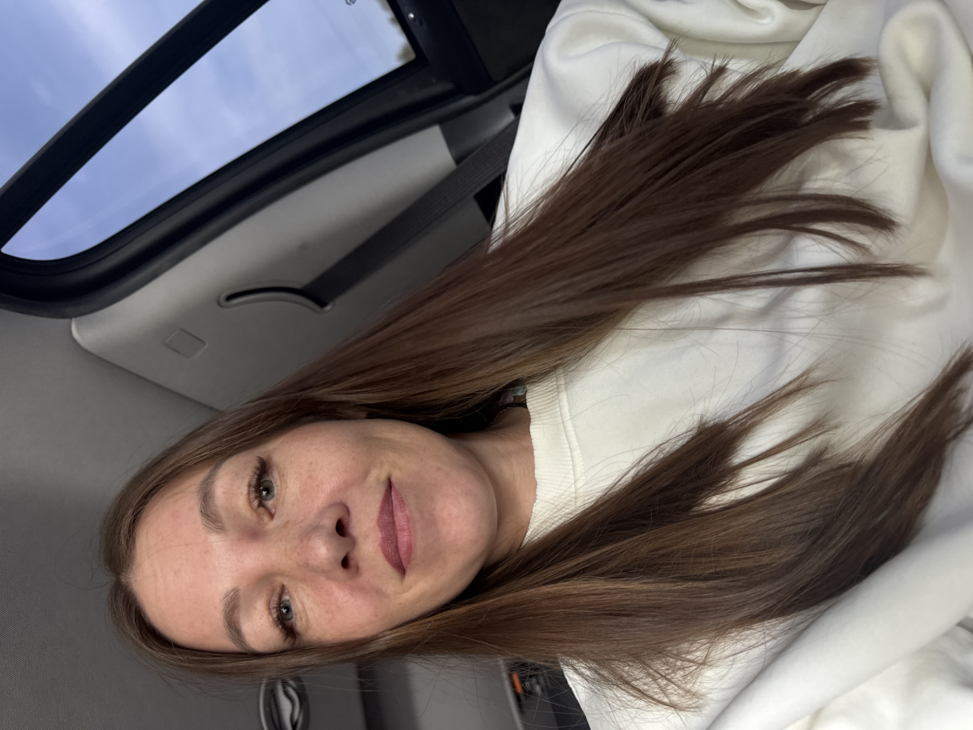 A woman with light skin and long brown hair is in a car. She is wearing a white sweatshirt. The car appears to have a sunroof. The location is unknown, but not confirmed to be in the city of Можга, Russia. There are no other people, objects, actions, or interactions directly visible in the image.