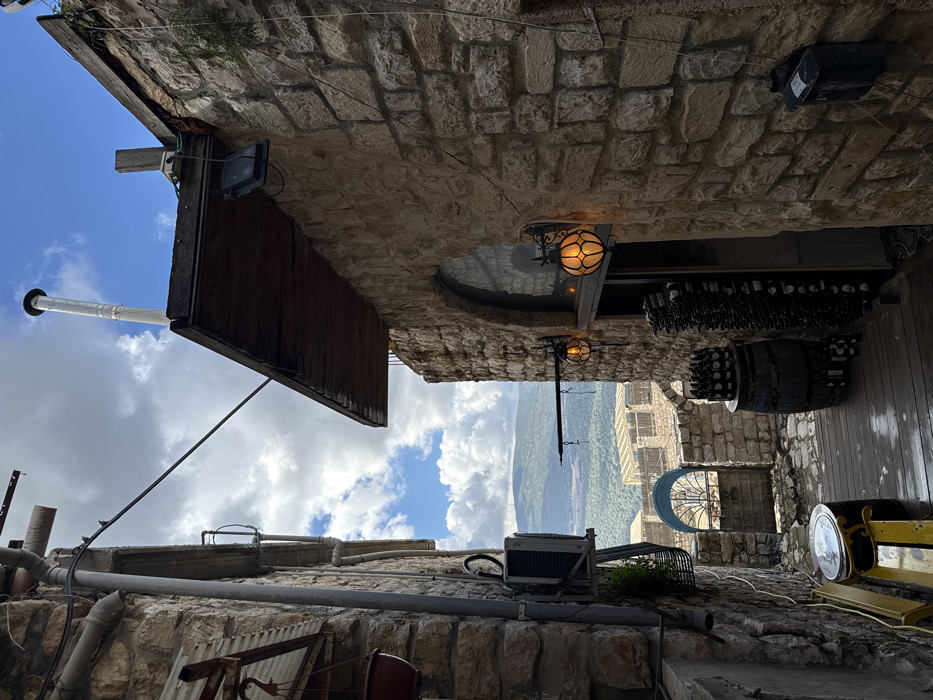 This image depicts a narrow stone alleyway located in Safed, Israel, under a partly cloudy sky with visible blue patches.
The left side of the alley is defined by a multi-story building constructed from rough-hewn stone blocks. This wall features external utility pipes, an air conditioning unit with a visible fan, and a red hanging planter. A dark cable extends diagonally across the upper portion of the frame.
The right side of the alley is bordered by another multi-story building, also constructed from rough-hewn stone. A wooden awning projects horizontally from this structure. Below the awning, two ornate, amber-glowing globe-shaped wall lanterns are mounted, flanking an arched opening or window. A black, rectangular outdoor light fixture is also affixed beneath the awning. A slender, metallic chimney pipe extends vertically from the building's rooftop. A dark, rectangular box, possibly a mailbox or utility box, is attached to the lower right portion of this wall.
The ground surface of the alley consists of wet wooden planks in the foreground, transitioning to irregular stone paving further back. On the right side, a wooden barrel stands upright, topped with several dark-colored bottles. Adjacent to the barrel, a tall, vertical display rack holds numerous identical bottles, indicative of a wine display. On the left side of the wooden planks, a yellow wooden bench is present, with a dark, cylindrical object placed upon it.
The alley opens up to a panoramic view of a green valley with distant hills or mountains under the partly cloudy sky. In the mid-ground, a lower stone structure featuring an arched blue gate and a building with a slatted roof are visible before the distant landscape. No individuals are present within the scene.