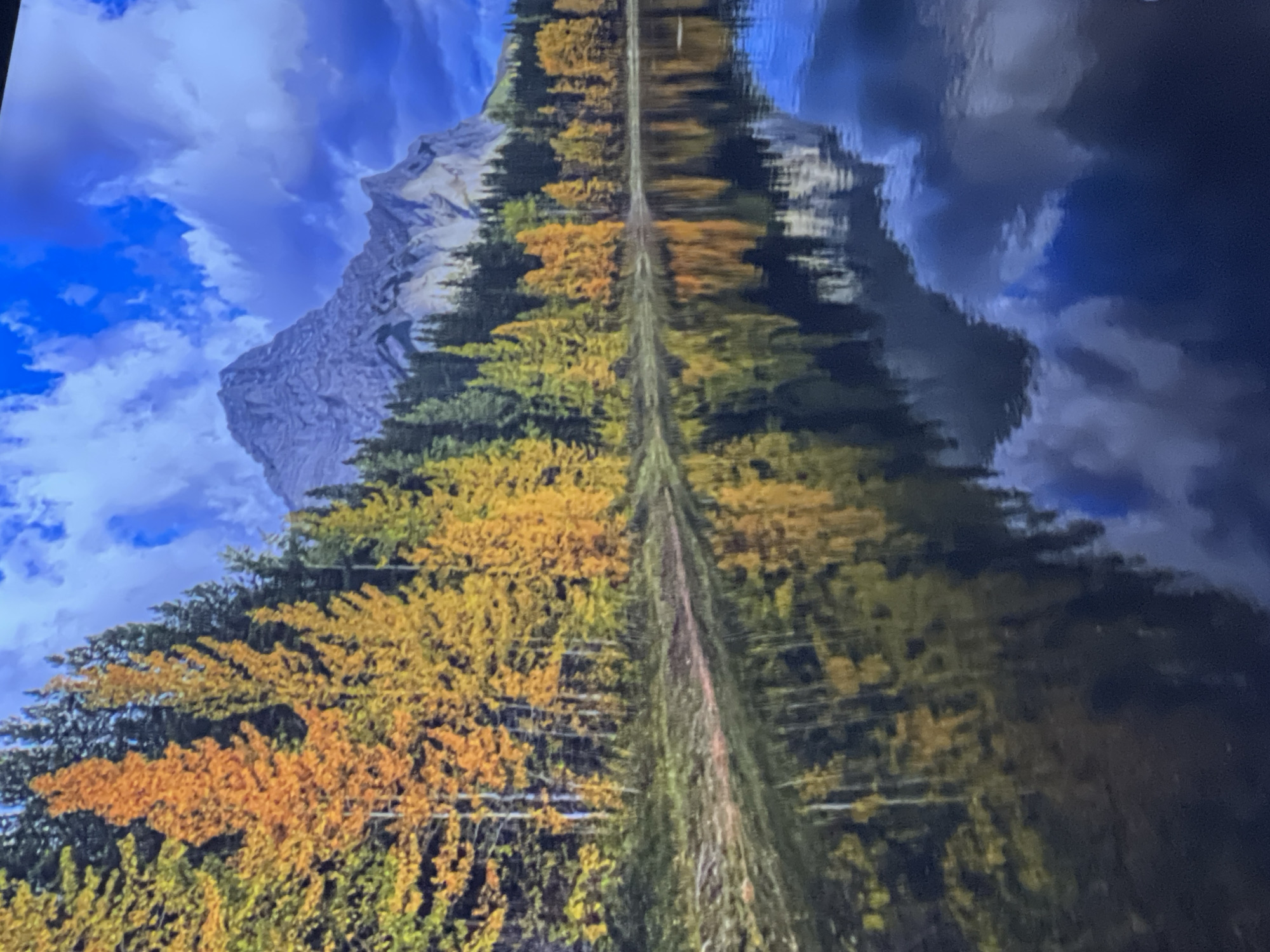 The image depicts a serene natural landscape. In the foreground, a calm body of water perfectly mirrors the scene above. The water's surface reflects a shoreline lined with trees displaying vibrant autumn foliage in shades of yellow and orange, interspersed with dark green evergreen trees. Beyond the trees, a majestic, rocky mountain peak rises, its rugged contours softened by the distance. The sky above is a dramatic mix of bright blue expanses and voluminous, dark clouds, adding depth and contrast to the scene. The reflection in the water captures the colors of the trees and the dramatic sky, creating a symmetrical and tranquil visual. There are no people, structures, or any indication of the city of Odesa, Ukraine in this image.