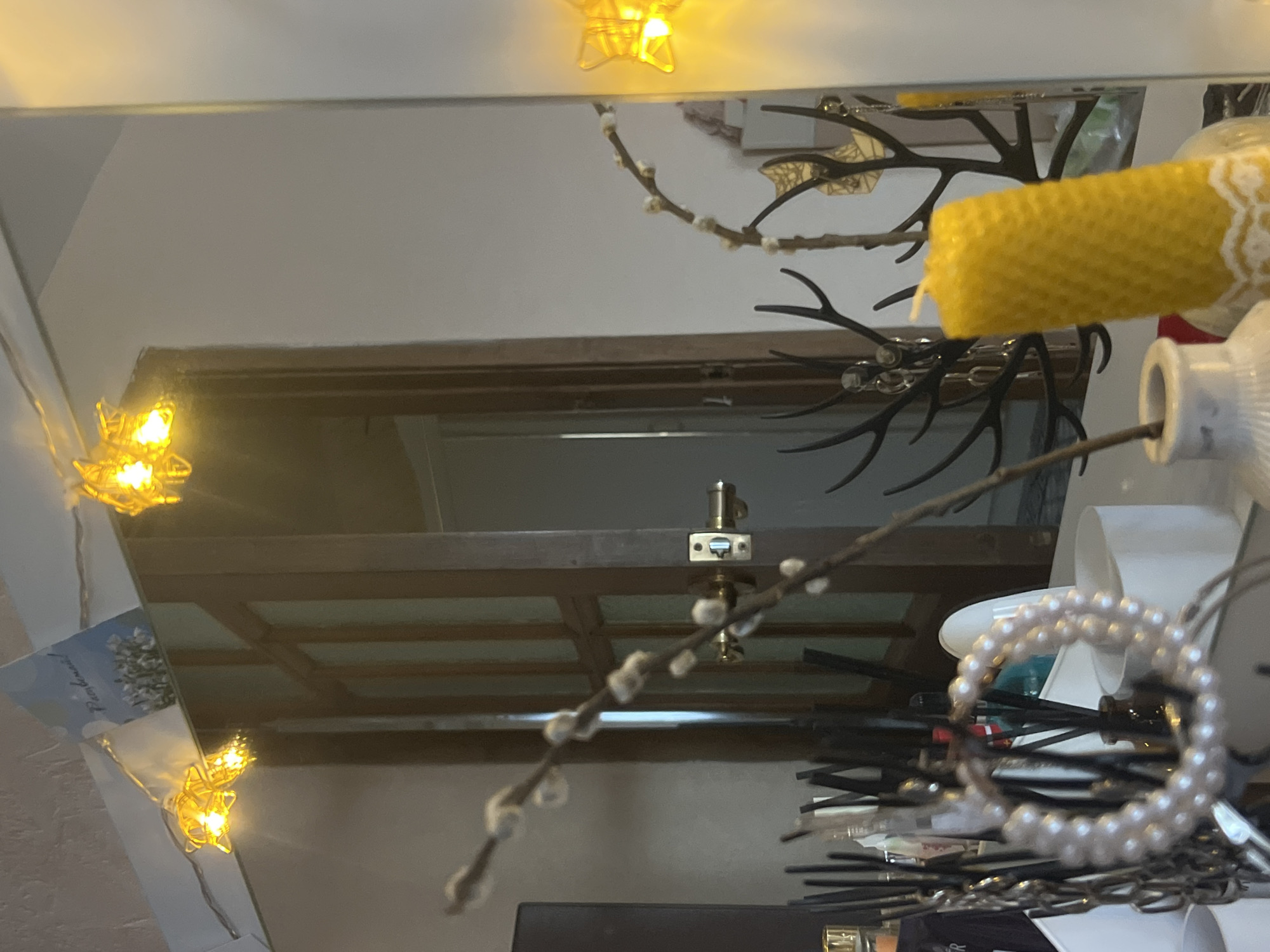 The image appears to be a reflection in a mirror, showing a window with a latch, decorative items including a yellow candle, artificial antlers, a string of lights, a beaded bracelet, and pussy willow branches. The reflection suggests an indoor setting, possibly a home. There is no information in the image to determine a location in Votkinsk, Russia.