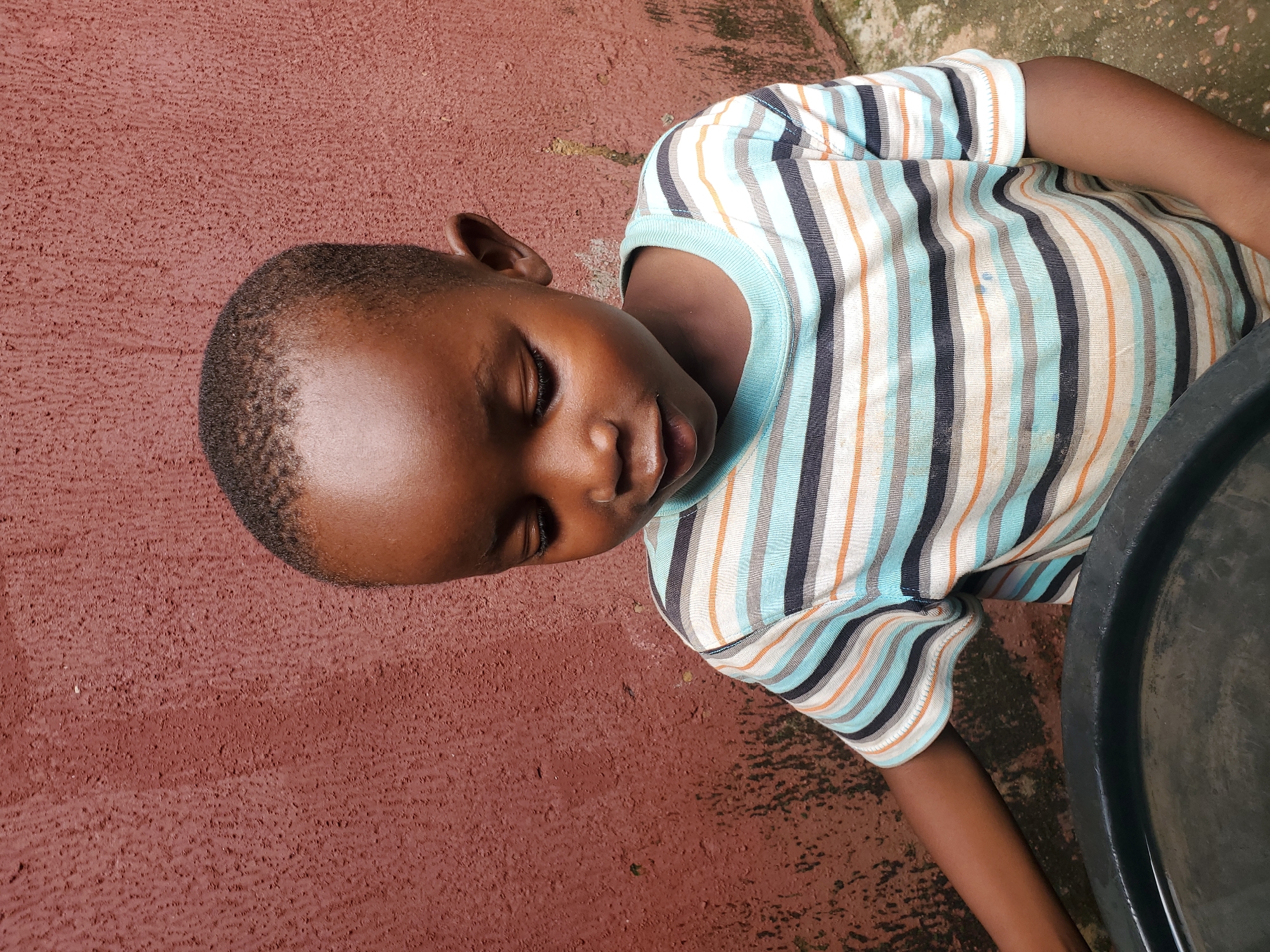 The media file shows a close-up, overhead view of a young Black child lying on their side on a reddish-brown, textured surface. The child is wearing a striped t-shirt with vertical bands of blue, white, black, and orange. The child's eyes are closed, and their head is resting on the surface. Their right arm is visible, extended downwards, and their left arm is not fully in view. To the right of the child, a portion of a large, dark, circular object, possibly a basin or container, is visible. The ground appears to be outdoors, with some lighter patches that might indicate sunlight or moisture. The overall impression is of a moment of rest or sleep in an outdoor setting. There is no visible text in the image.