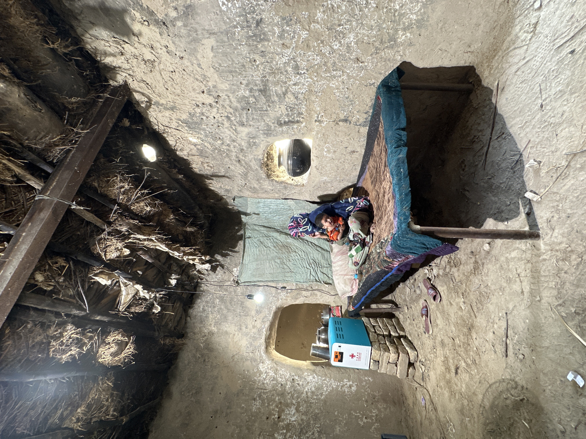 A person is lying on a cot in a dimly lit, rudimentary room. The walls and ceiling of the room are constructed of earth or clay. There are openings in the walls for light and ventilation. The ceiling appears to be supported by a structure of wood and straw. A light bulb illuminates the room. A medical device sits on the floor near the cot. Two sandals are visible near the bed. The location is Quetta City Tehsil, Pakistan.