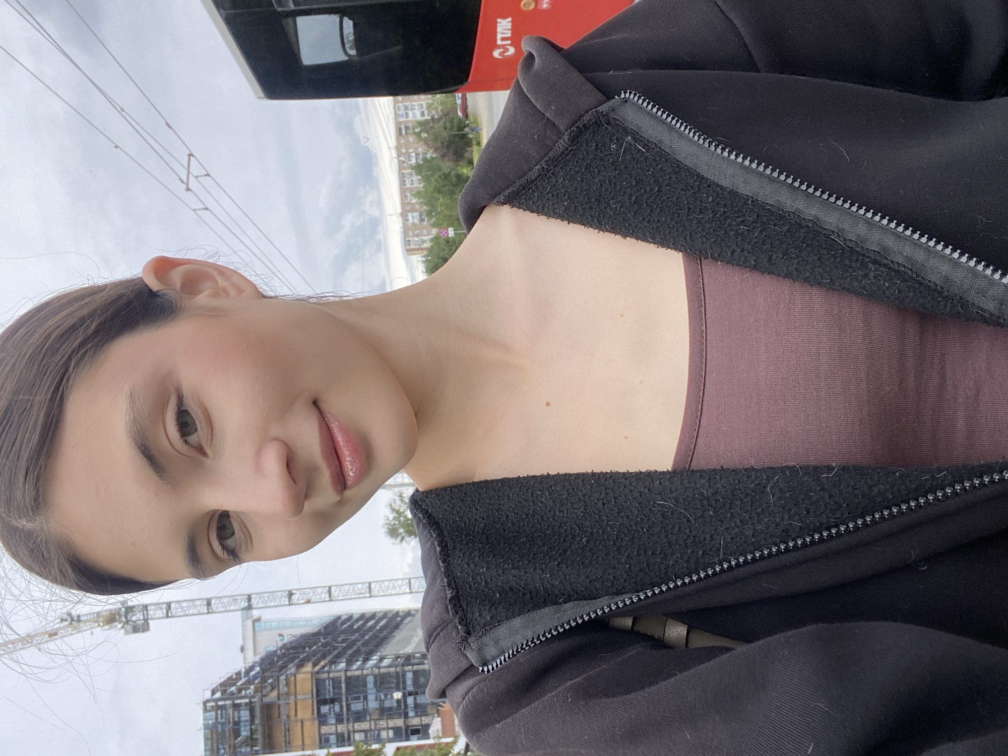 A young woman with brown hair pulled back and fair skin takes a selfie outdoors in Izhevsk, Russia. She is wearing a dark hooded jacket partially zipped over a rust-colored tank top. In the background, a construction crane and an unfinished building are visible on the left, with power lines overhead. To the right, the rear of a red public bus with the text "ГЛПК" and Cyrillic lettering is partially visible. The sky is overcast with clouds.