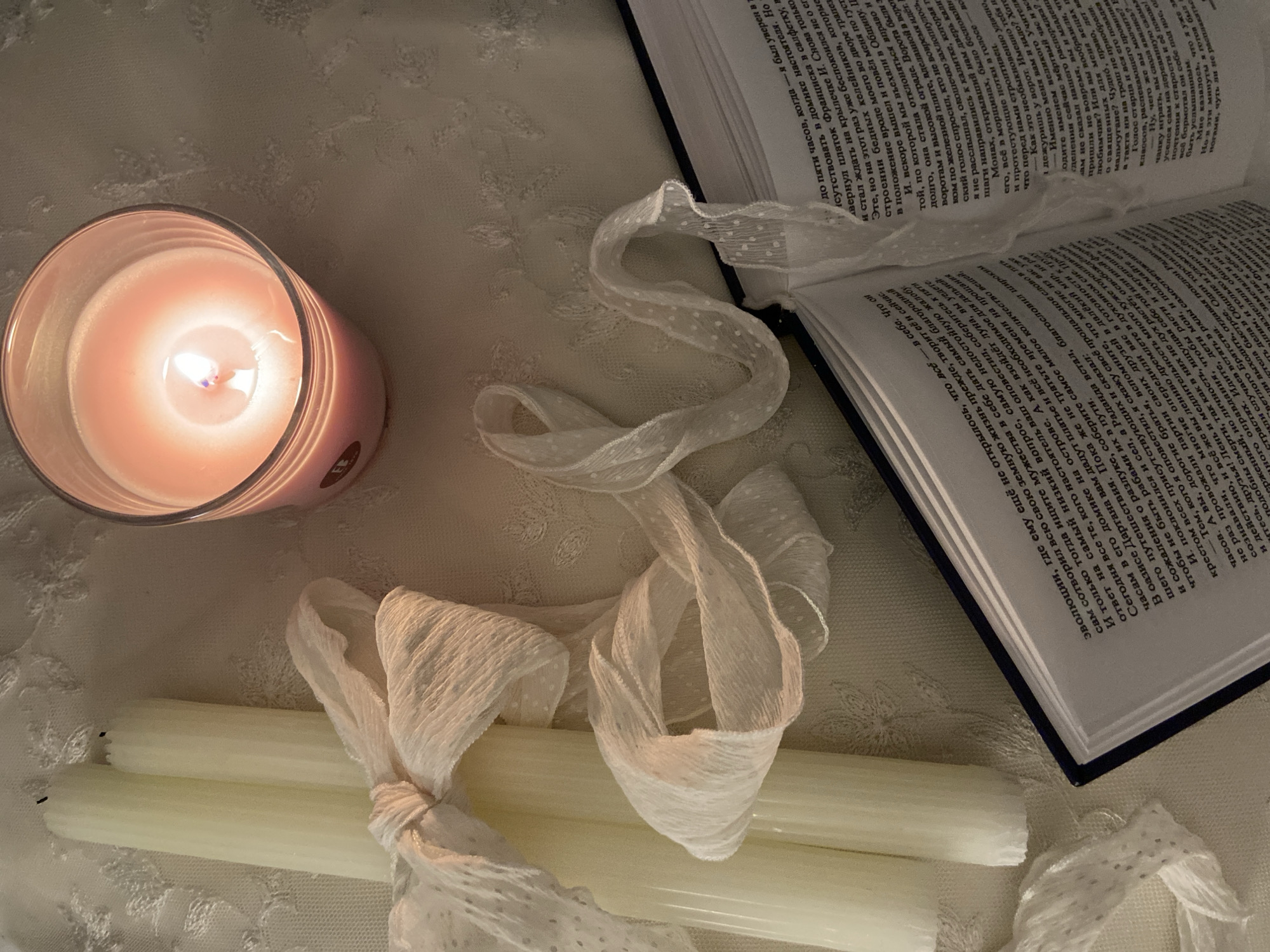 The image presents a still life arrangement on a soft, textured surface, likely a tablecloth. A lit pink candle sits to the left, casting a warm glow. Next to it, two unlit white candles are tied together with a sheer, dotted ribbon. The ribbon continues, draped loosely around the arrangement. An open book with dense text lies to the right, its pages angled towards the viewer. The lighting is soft, creating a cozy and intimate atmosphere.