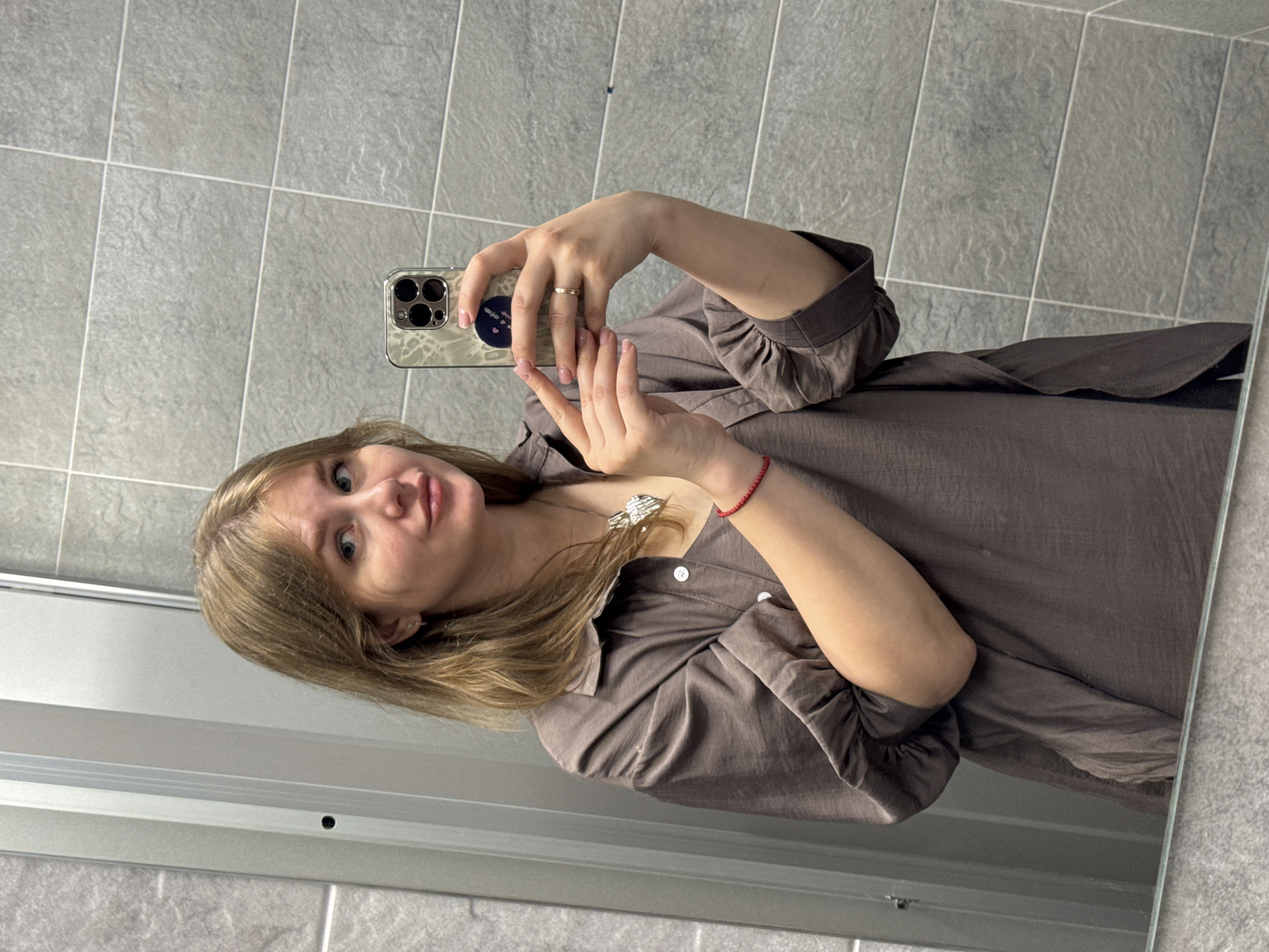 A young woman takes a selfie in a bathroom. She has blonde hair and is wearing a brown button-down shirt. She holds an iPhone with a sticker on the back in her right hand. The bathroom features gray tile walls and a mirror with a metal frame. This scene is likely in Perm, Russia.