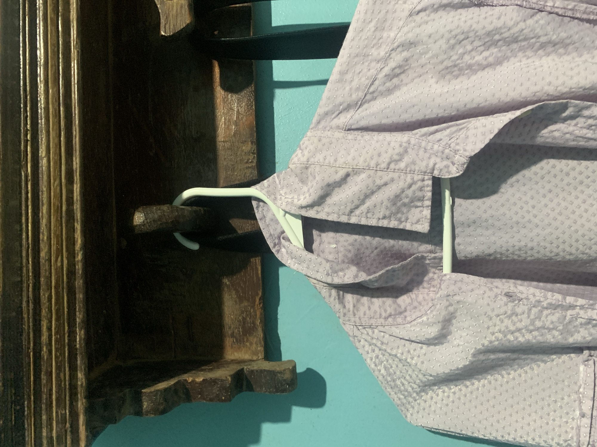 The image captures an indoor scene, primarily featuring a light purple, long-sleeved shirt with a subtle textured pattern of small, raised dots or squares. The shirt is hanging on a white plastic hanger and occupies the right side of the frame. The visible portion includes the collar and upper body of the garment.
A bright turquoise wall forms the background. To the left, a dark, ornate wooden structure, possibly a shelf or a furniture piece, features vertical fluting on its left side and decorative carvings near its base. A black strap or belt is draped behind the hanger, resting against the wooden structure and the wall.
Shadows from the shirt, hanger, and wooden structure are cast on the turquoise wall, indicating an indoor light source. No people or visible text are present in the image. The time of day and weather conditions are not discernible. The provided location context is Santa Ana, Venezuela, though no specific visual cues within the image confirm this.