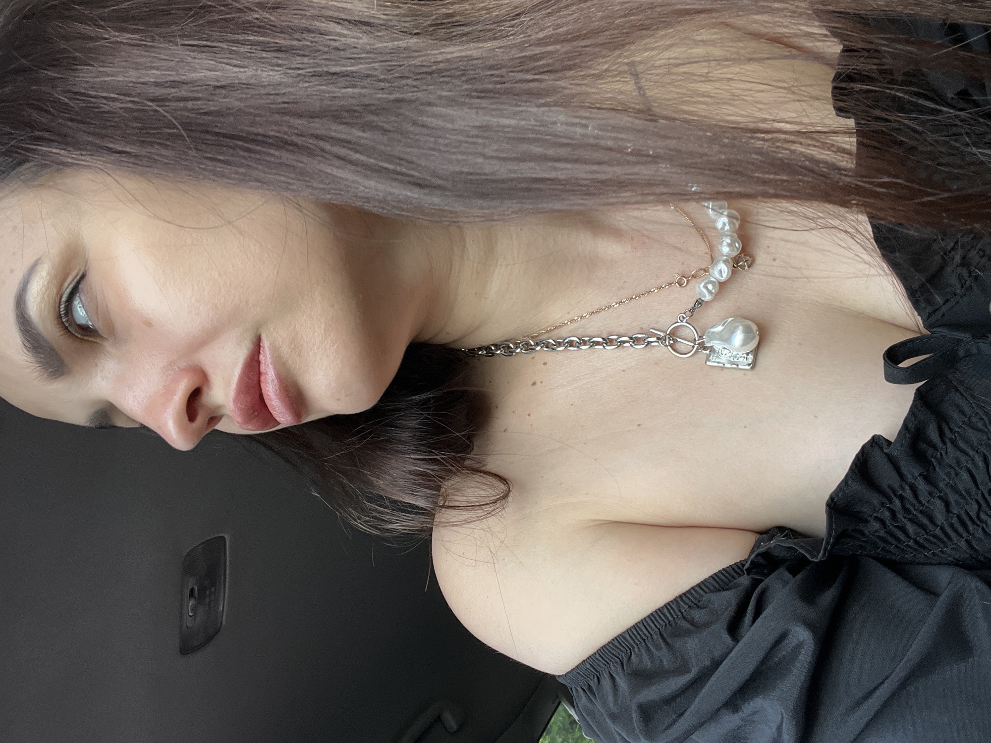 A close-up, angled portrait features a person, likely a woman, with long, dark brown hair, fair skin, and subtle makeup, including winged eyeliner and soft pink lipstick. Her head is tilted, and her gaze is directed upwards and slightly to the left. She is wearing a black off-the-shoulder top or dress, characterized by shirring or elasticated fabric along the neckline and sleeves.
Adorning her neck are layered necklaces. One is a prominent silver chain with a toggle clasp, featuring a unique baroque pearl pendant. Another appears to be a delicate rose gold chain, potentially combined with a strand of small, round pearls.
The background is dark and out of focus, suggesting the interior of a vehicle, possibly a car, with a dark panel or handle visible on the left. The soft, even lighting indicates daytime, but without direct sunlight. The person appears to be posing for the camera, exhibiting a calm and neutral expression. There is no visible text in the image.