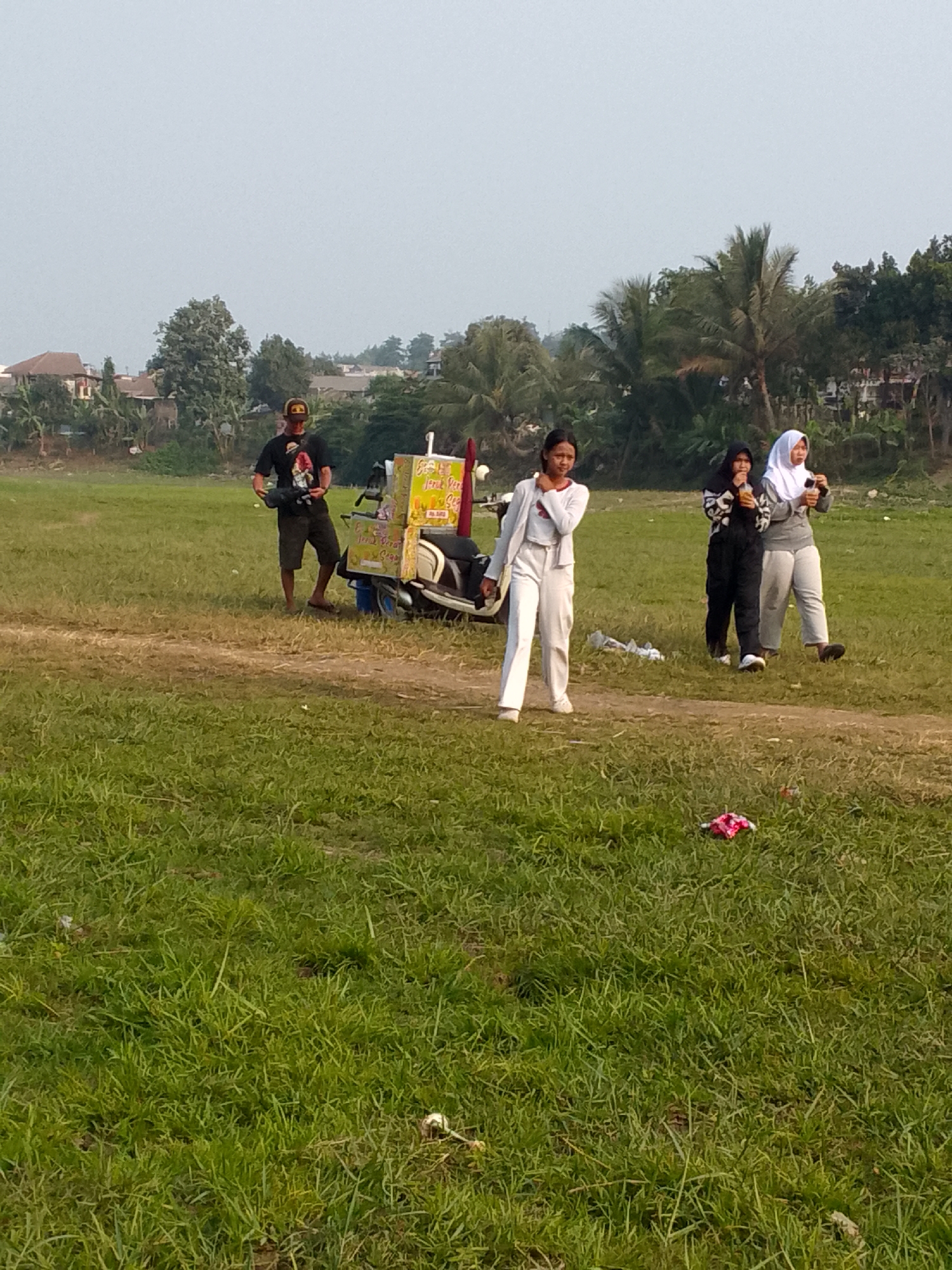 A wide, low-angle shot captures an outdoor scene in Ciburuy, Indonesia, featuring a grassy field with a dirt path running through it. In the middle ground, a man wearing a black t-shirt with a graphic print, khaki shorts, and a baseball cap stands near a three-wheeled cart equipped with a yellow canopy and signage. He appears to be holding something in his hands. To his right, three young women are present. The first woman, on the far left, wears a white blazer over a white top and white pants. The second woman, in the center, is dressed in a black patterned top and black pants, holding a yellow cup. The third woman, on the right, wears a white hijab and a gray top with white pants, also holding a yellow cup. Behind the individuals and cart, a line of trees and residential buildings forms the backdrop under a hazy sky. In the foreground, scattered across the grass, are some bits of litter, including a cluster of pink and red items.