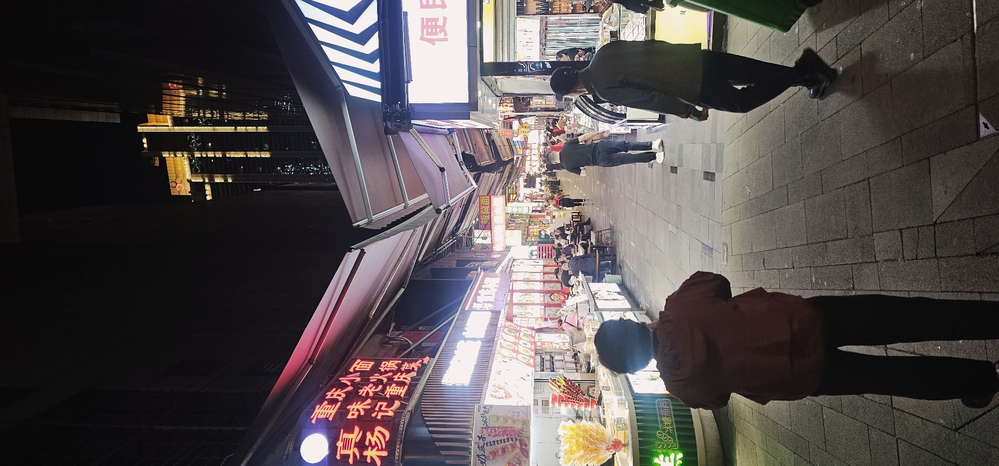The image shows a street scene in Yuzhong District, Chongqing, China, at night. The view is along a pedestrian street lined with shops and food stalls. Several people are walking along the street. Many of the stalls have bright signs and displays of food. Buildings are visible in the distance.