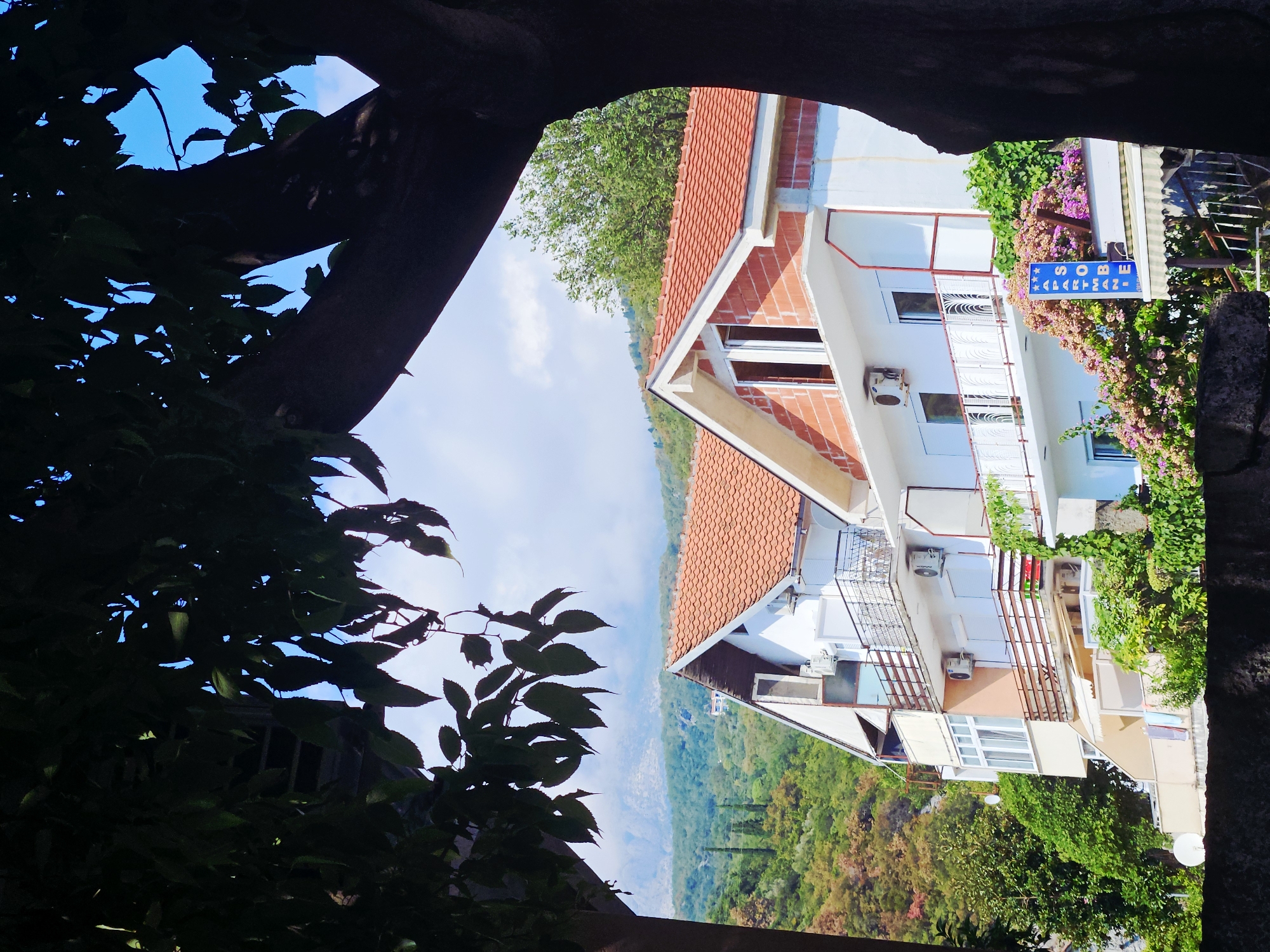 A view of a building with tiled roofs and balconies is framed by the dark branches and leaves of a tree in the foreground. The building is white with some red brick accents and has multiple windows and air conditioning units visible. A sign with blue stars and the word "SOBE" is mounted on the building, along with another sign that reads "APARTMANI". Lush green trees and vegetation surround the building, and in the background, there are rolling hills covered in trees and a hazy mountain range under a bright blue sky with scattered clouds.