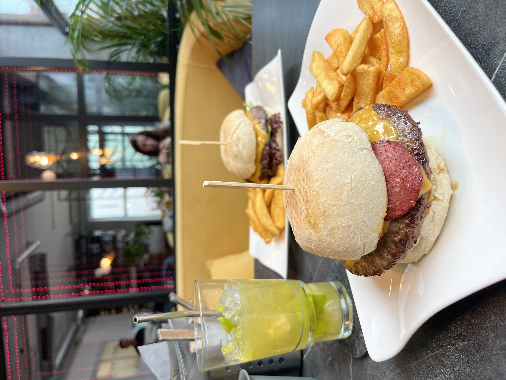 The photograph depicts a food and drink setting, likely in a restaurant in Bad Kreuznach, Germany. Two white plates are visible, each containing a burger and a side of thick-cut fries. The burger in the foreground has a patty, cheese, and what appears to be a slice of sausage between the bun. Both burgers are secured with a wooden toothpick. In front of the burgers is a glass of what looks like a lime-based cocktail with ice and a metal straw.
In the blurred background, there is a glimpse of a seating area with a yellow sofa and an individual seated. A window with red decorative lights surrounds the window frame is also visible. There is also a glimpse of what appears to be a palm tree to the left. The surface the plates and drink rest on is dark and has a speckled appearance.