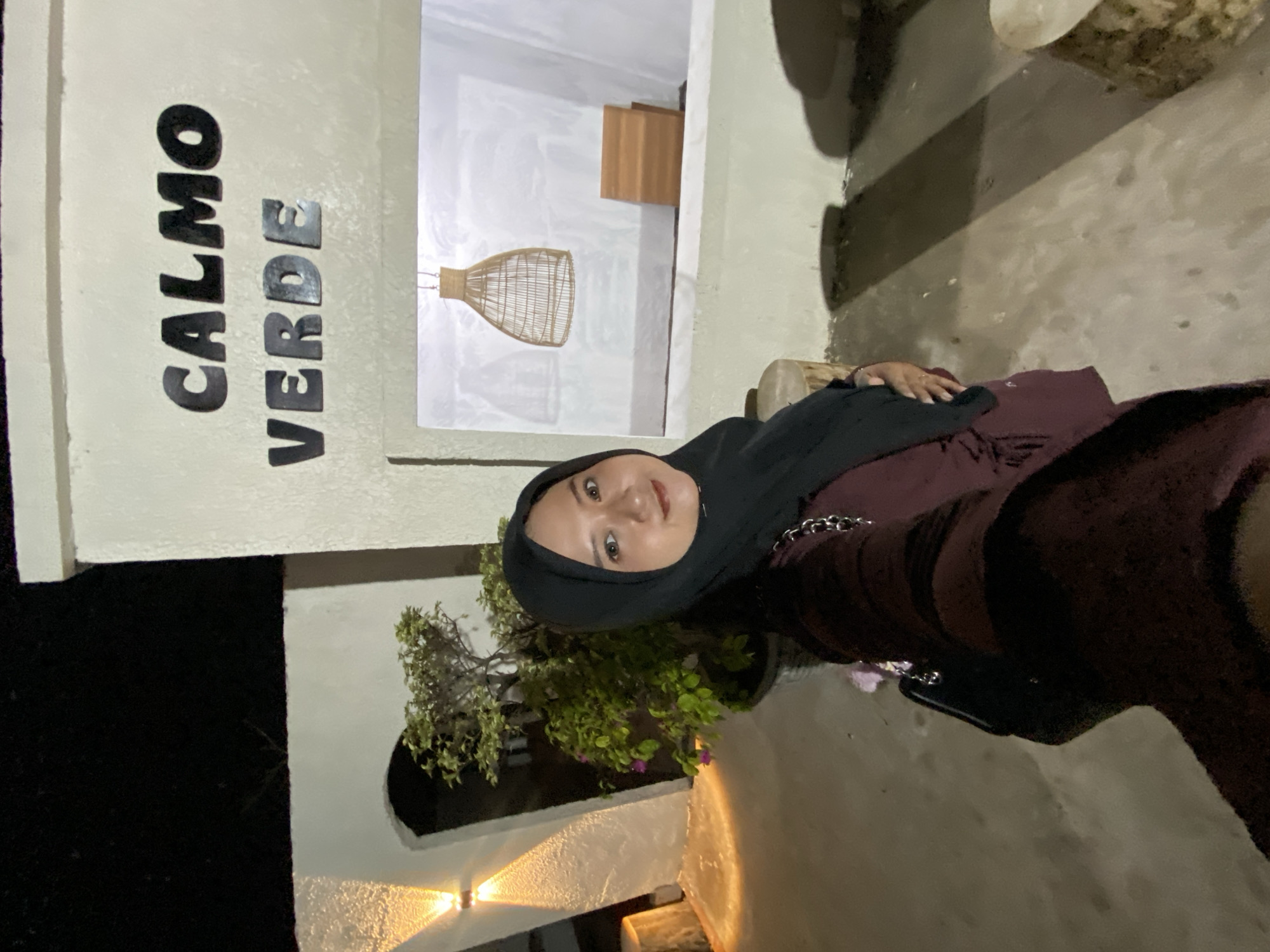 A medium shot captures a woman in a black hijab and maroon dress posing for a selfie in front of a white stucco building. The building features bold, dark lettering on its facade that reads "CALMO VERDE" in two lines. To the left of the woman, an arched doorway is illuminated by two upward-facing lights, and a potted plant with green foliage and small pink flowers sits within the arch. To the right, a rectangular opening reveals a rustic woven pendant light fixture hanging from the ceiling and a small wooden shelf with two horizontal planks. The ground is paved with concrete. The scene is set at night, indicated by the dark sky in the upper portion of the image and the artificial lighting.